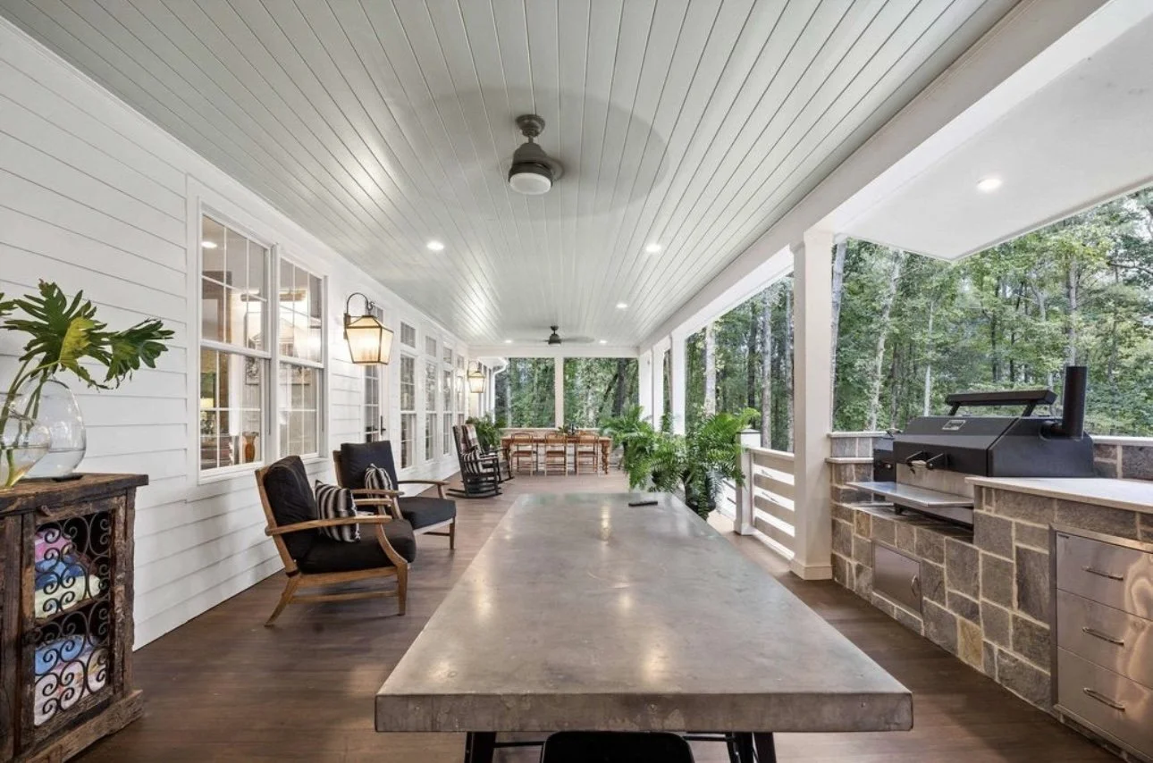 Covered porch area with a concrete table, black chairs, potted plants, and a built-in grill, overlooking a wooded backyard.