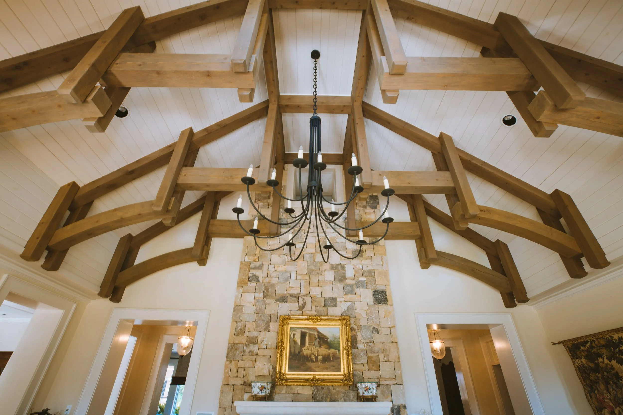 Interior of a room with a high vaulted ceiling made of light wood beams and panels, a large black chandelier hanging in the center, and a stone fireplace with a framed painting above it.