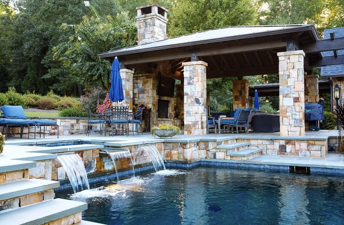 A backyard with a stone pool, water features, and an outdoor seating area under a pavilion supported by stone columns, surrounded by greenery.