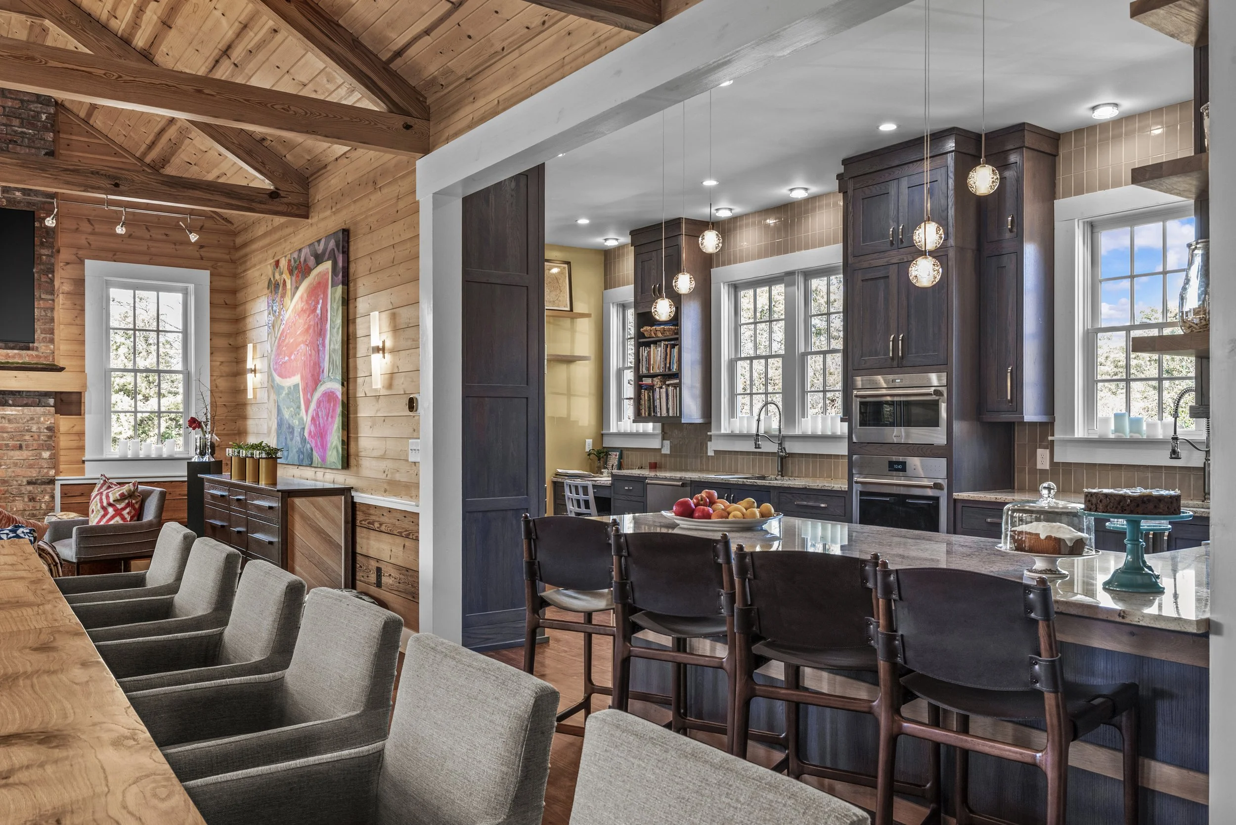 Open-concept kitchen and dining area with wooden and brick accents, dark wood cabinetry, large windows, and hanging pendant lights, decorated with apples and cake on the counter.