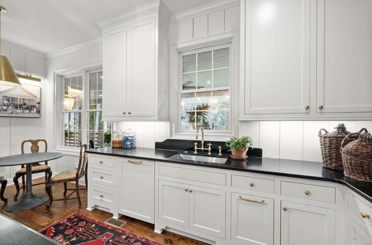 Kitchen with white cabinets and black countertop, window with ocean view, sink with brass faucet, potted plant, wicker baskets, wooden dining table and chairs, red patterned rug on wooden floor.