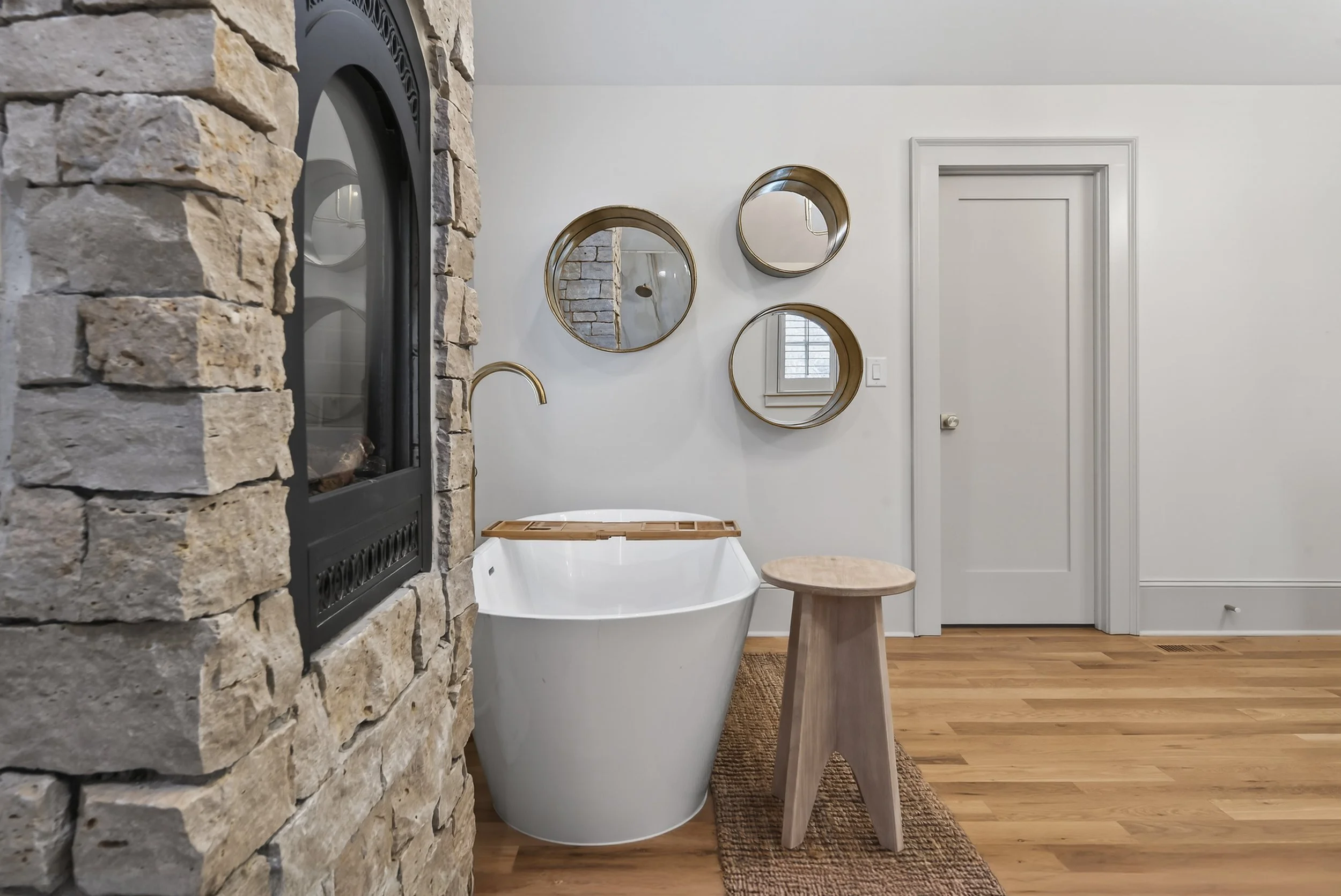 A modern bathroom with a stone wall fireplace on the left, a white bathtub with a wooden tray lid, a small wooden stool, and four round mirrors on the wall. There is a window in the reflection of the mirrors and a wood floor with a small rug.