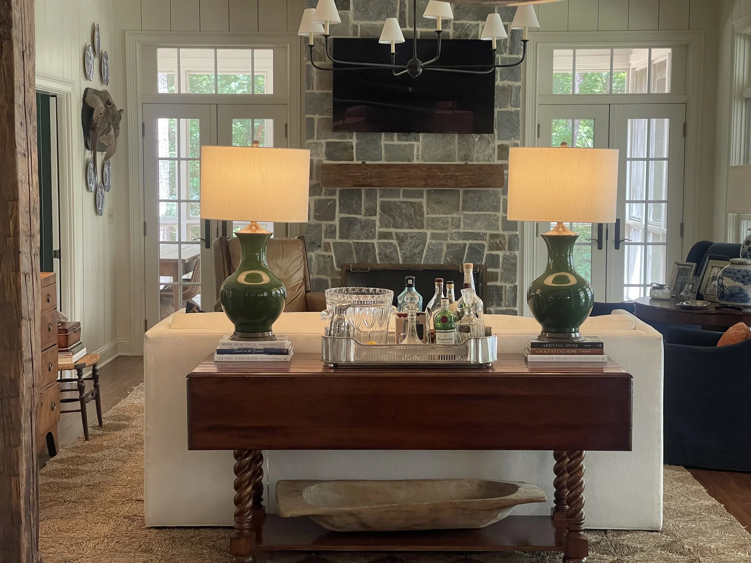 Living room with stone fireplace, wooden mantle, two large green lamps on a wooden console table, sofas, and glass doors leading outside.
