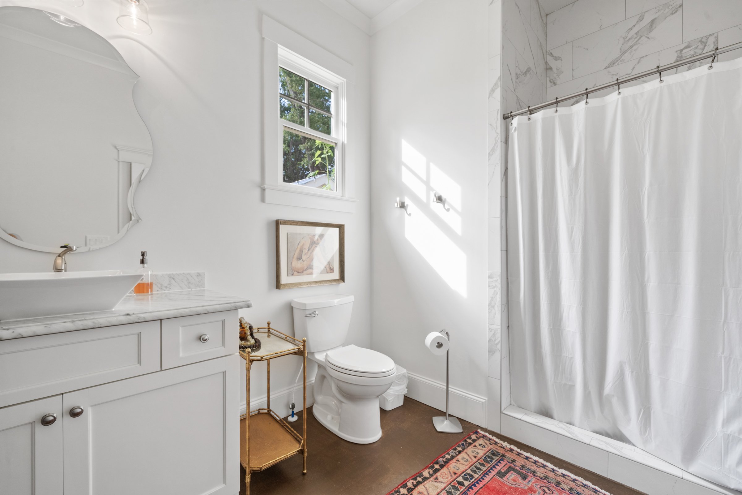 Bright white bathroom with a window letting in natural light, a white vanity with a marble countertop, a vessel sink, a mirror, a framed picture above the toilet, a gold stand, a toilet, a toilet paper holder, a white shower curtain, a colorful rug, 