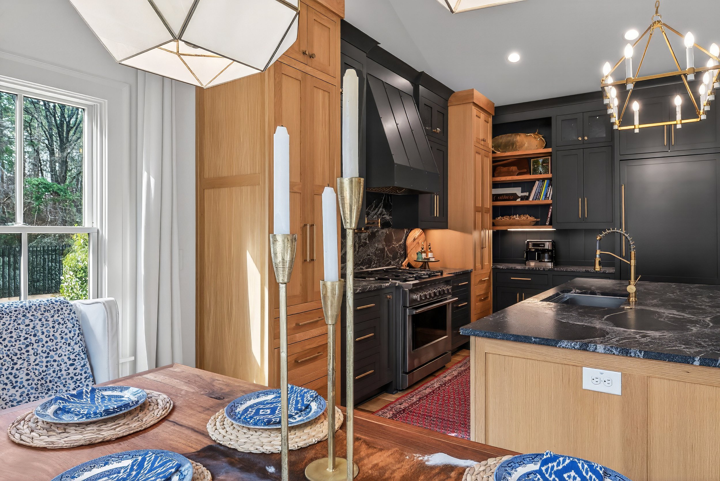 Modern kitchen with black and natural wood cabinetry, black marble countertop, and gold fixtures. Dining table with blue and white dishes, gold candle holders, and a large window with white curtains.