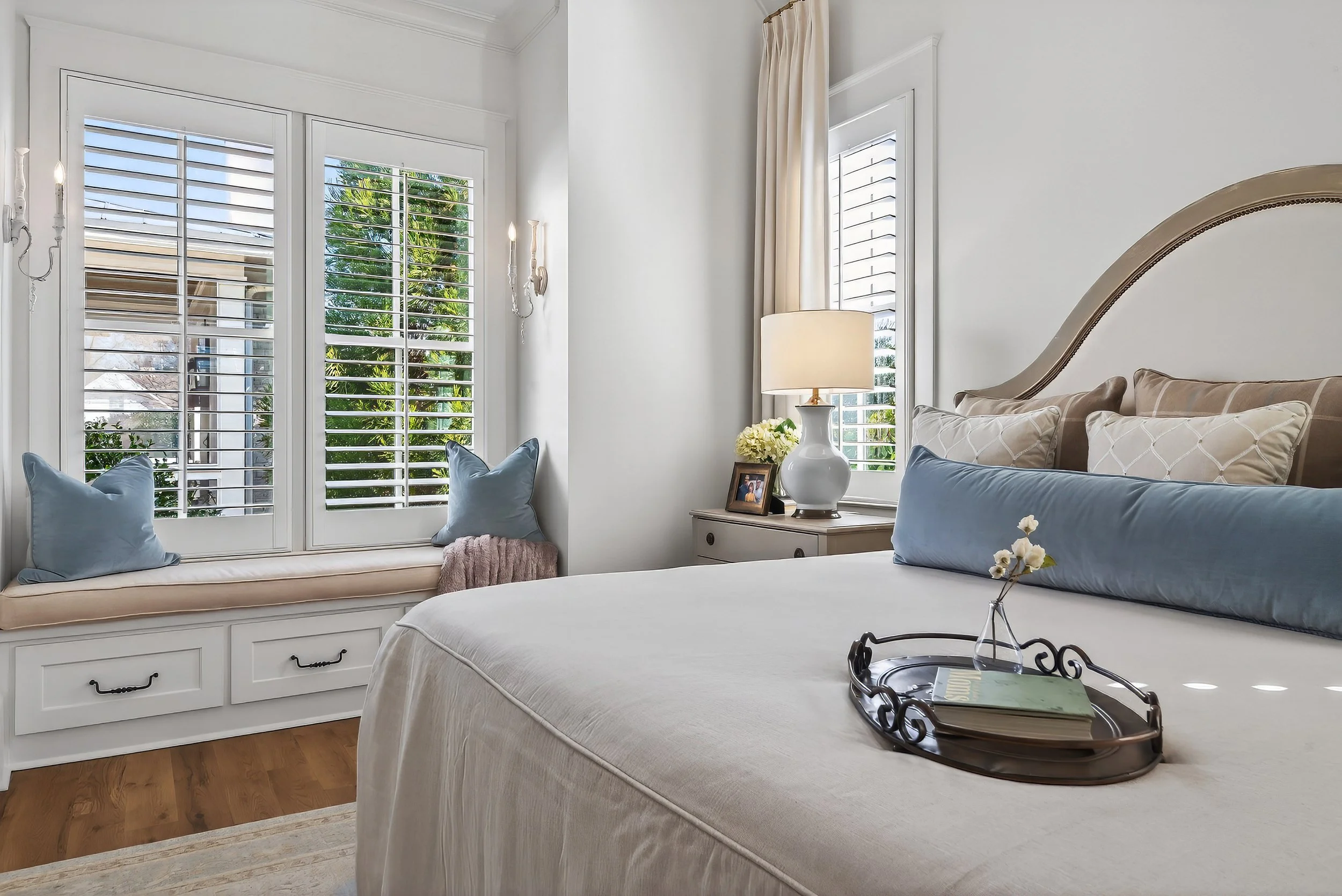 Bright bedroom featuring a bed with beige and blue pillows, a bedside table with a white lamp, a framed photo, and a bunch of white flowers. Large windows with white shutters and an window seat with throw pillows. Light-colored walls and hardwood floor.