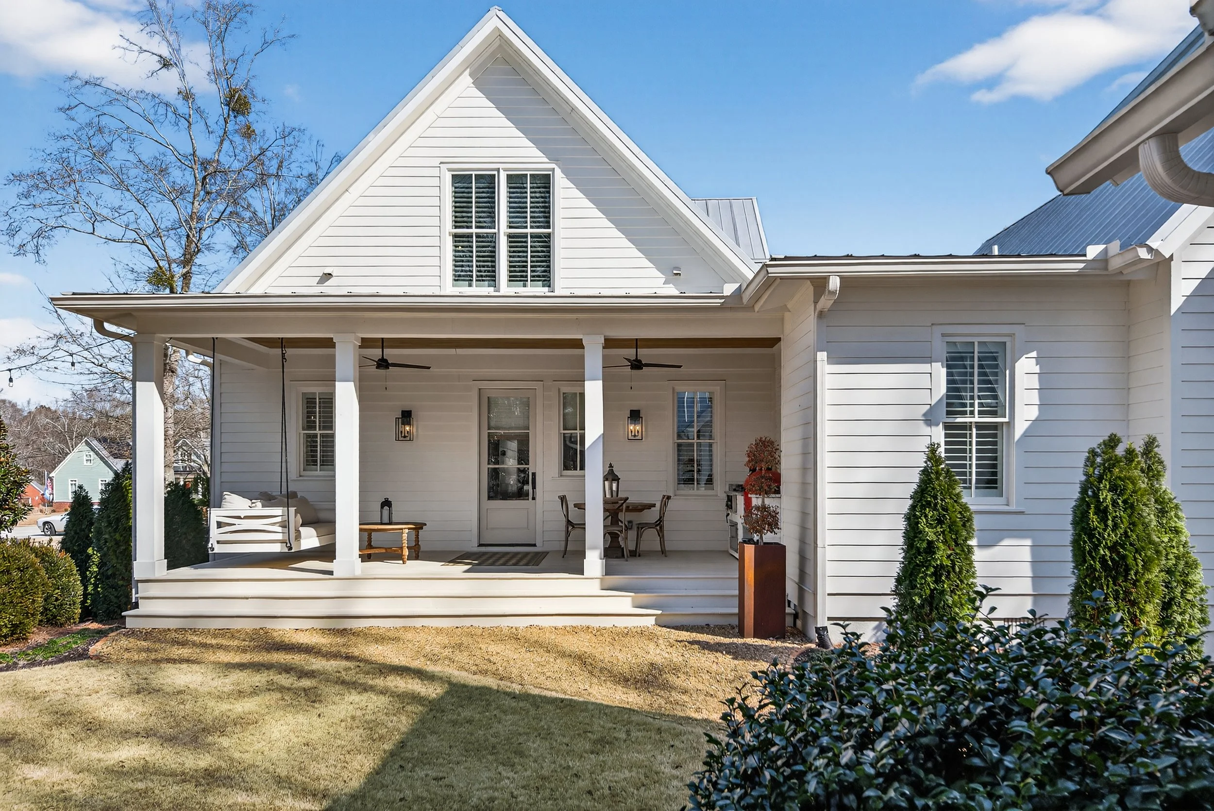 White house with a covered porch, two ceiling fans, and outdoor furniture, including a swing, table, and chairs, on a sunny day with a clear blue sky.