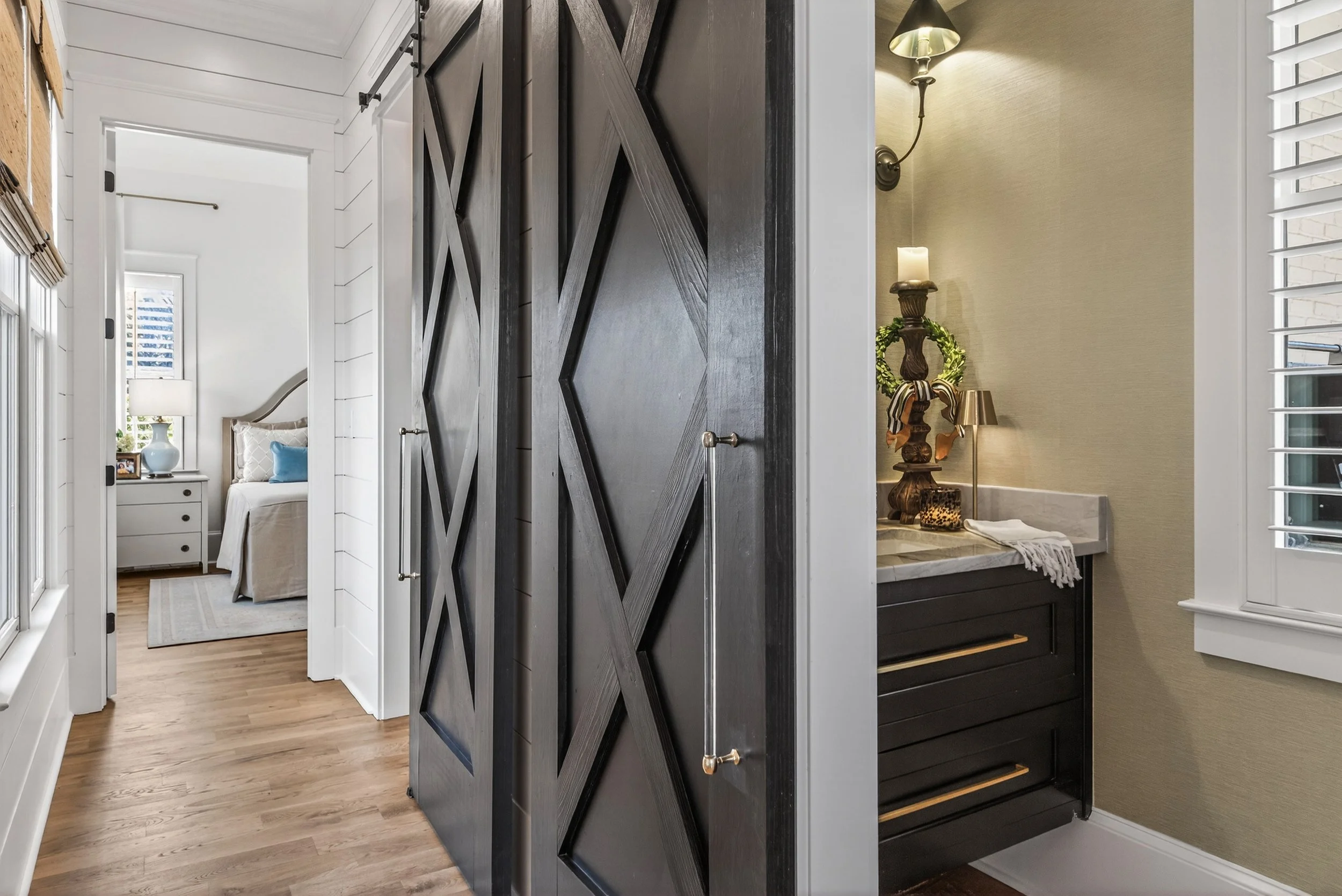 Interior view of a home hallway showing a bedroom with a white bed and nightstand on the left and a small bathroom with a black vanity, mirror, and decorative items on the right. The hallway has wooden floors and white walls and trim.