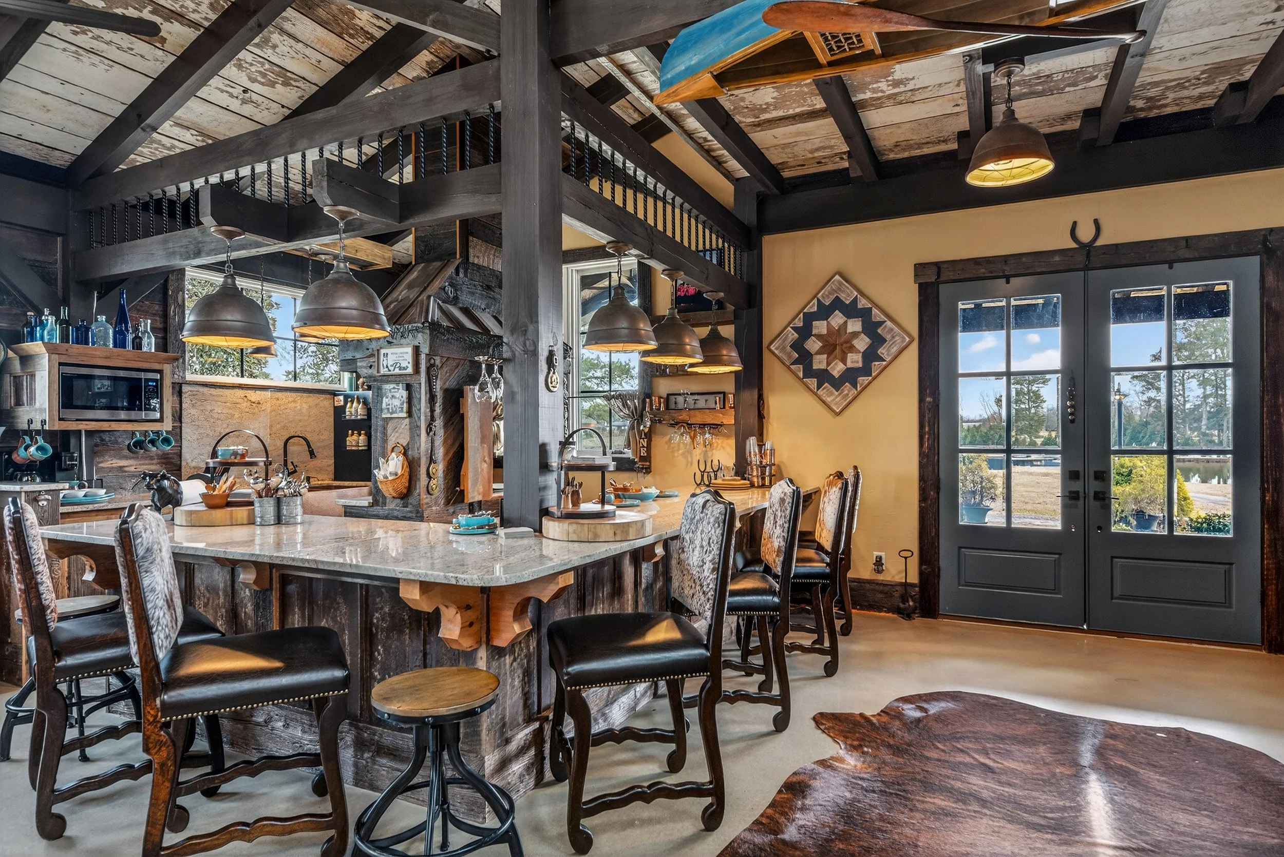 Rustic kitchen with a large marble island, black chairs, hanging pendant lights, wood accents, a door with glass panels showing an outdoor view, and a decorative quilt on the wall.