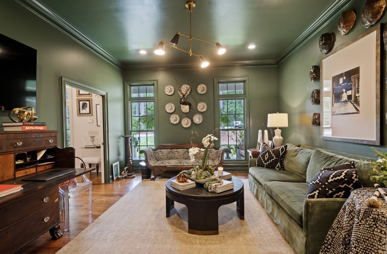 Living room with green walls, hardwood floors, a green sofa with decorative pillows, a round black coffee table with decor, and a bench with a floral pattern by the window. There's art and decorative plates on the walls, a ceiling light fixture, and 