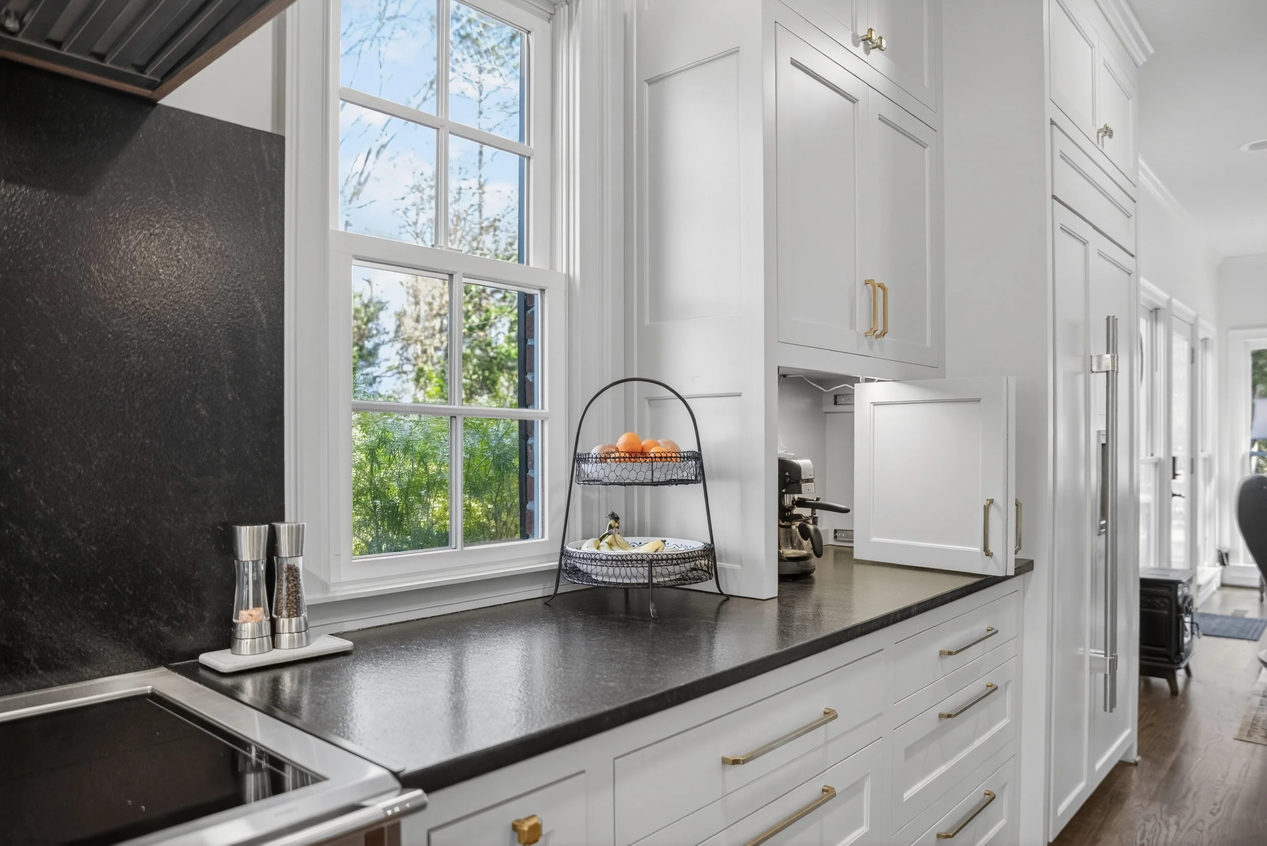 White kitchen with black countertop, window, fruit display, coffee machine, and white cabinets.