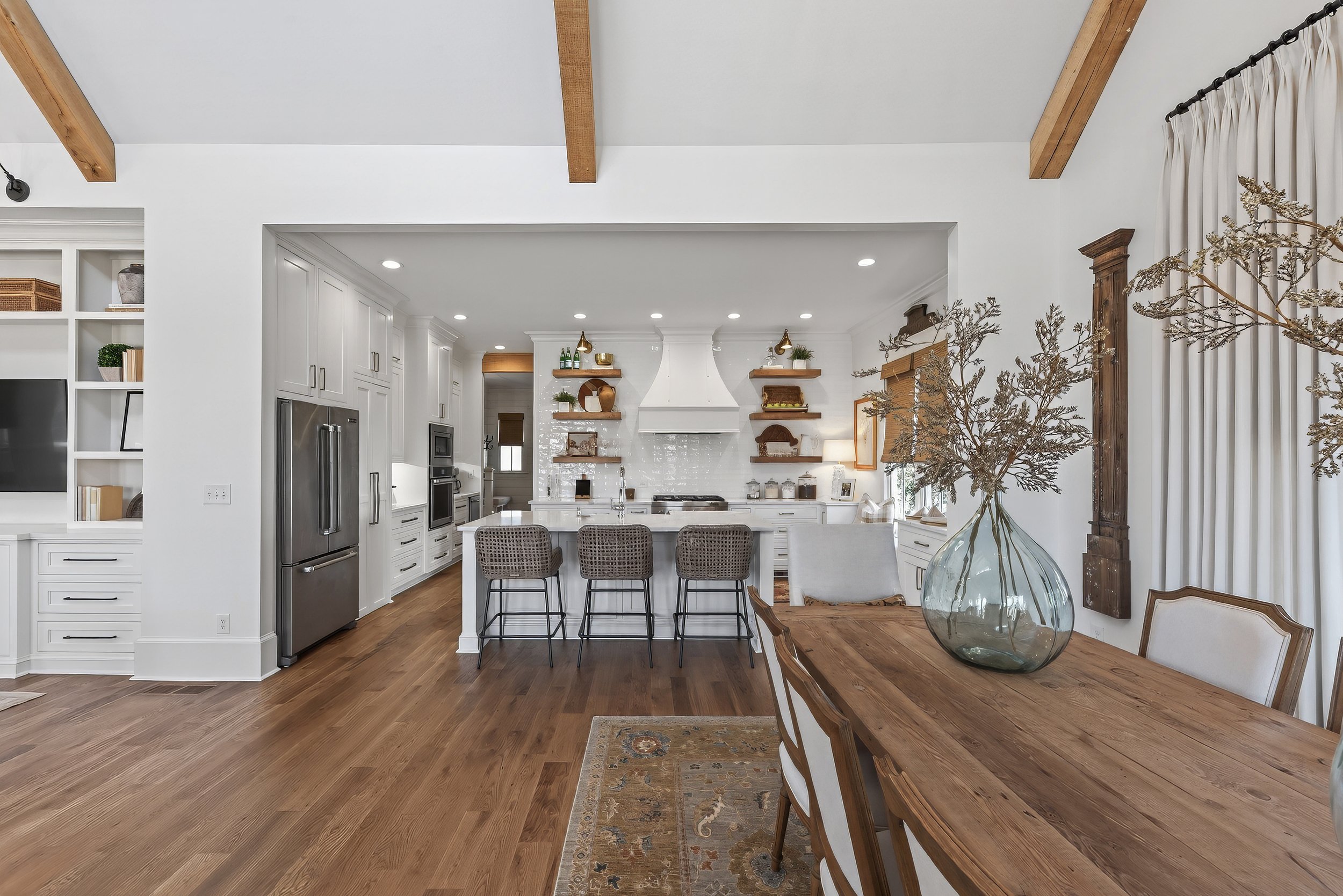 Open-concept kitchen and dining area with white cabinetry, stainless steel refrigerator, wooden floors, and a wooden dining table with chairs, decorated with a large glass vase of dried flowers.