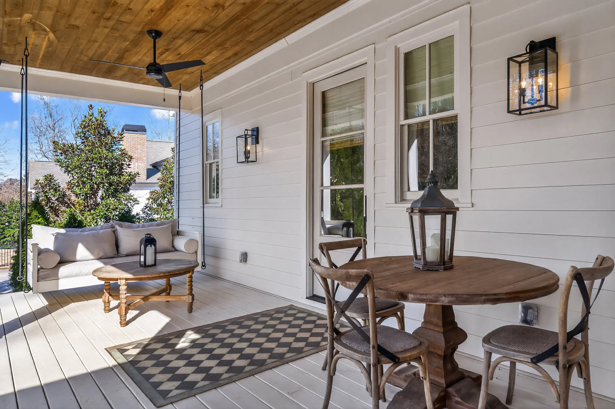 Covered porch with white siding, wooden ceiling, ceiling fan, black lantern-style wall lights, a white outdoor sofa with cushions, a wooden coffee table with lantern, a wooden round dining table with four chairs, a lantern on the table, a door, two w
