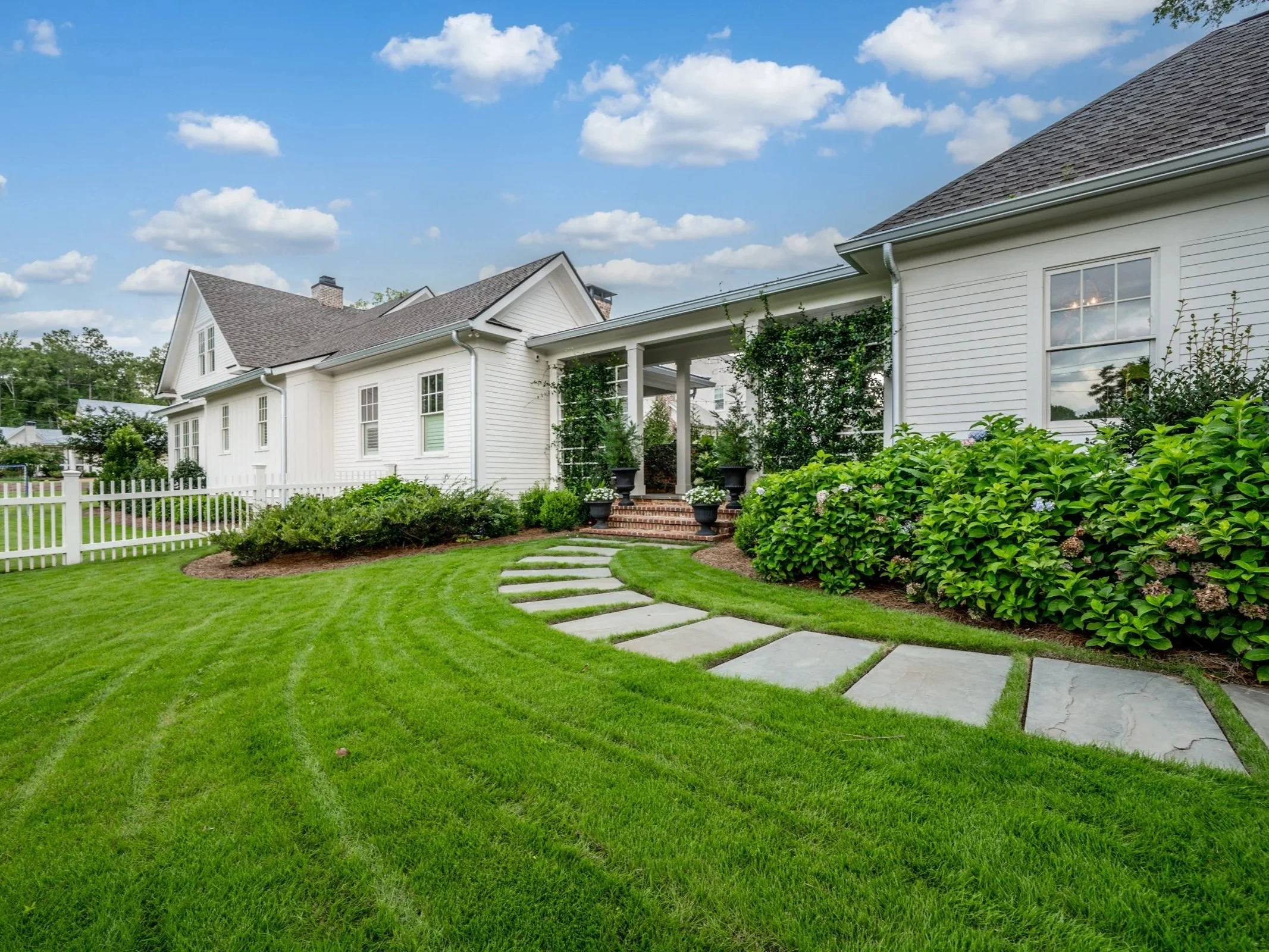 A well-maintained backyard with lush green grass, a curved stepping stone pathway, white picket fence, and a white house with large windows and a small porch with potted plants.