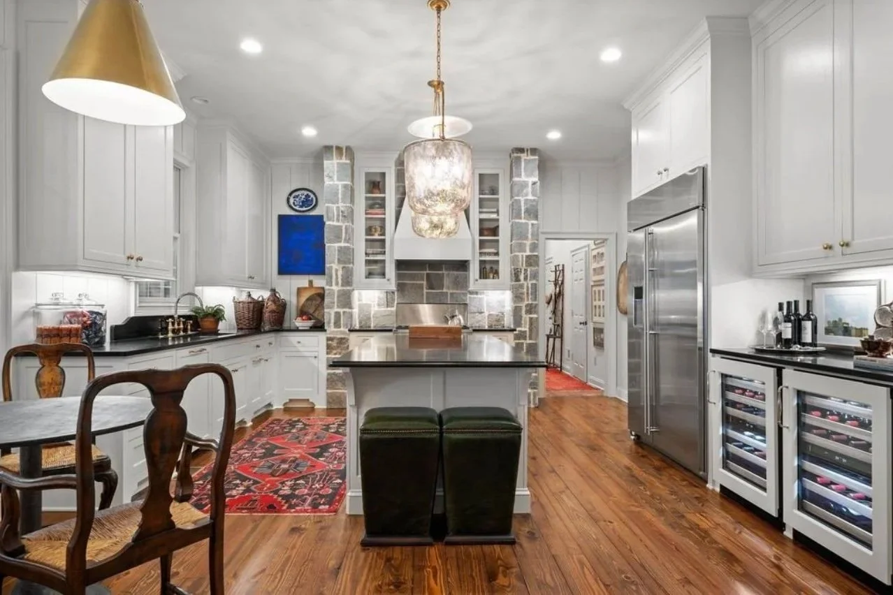 Modern kitchen with white cabinets, a central island with black countertop, a rustic stone fireplace with a chimney, hardwood floors, and decorative lighting. Contains wine coolers, a stainless steel refrigerator, and various kitchen accessories.