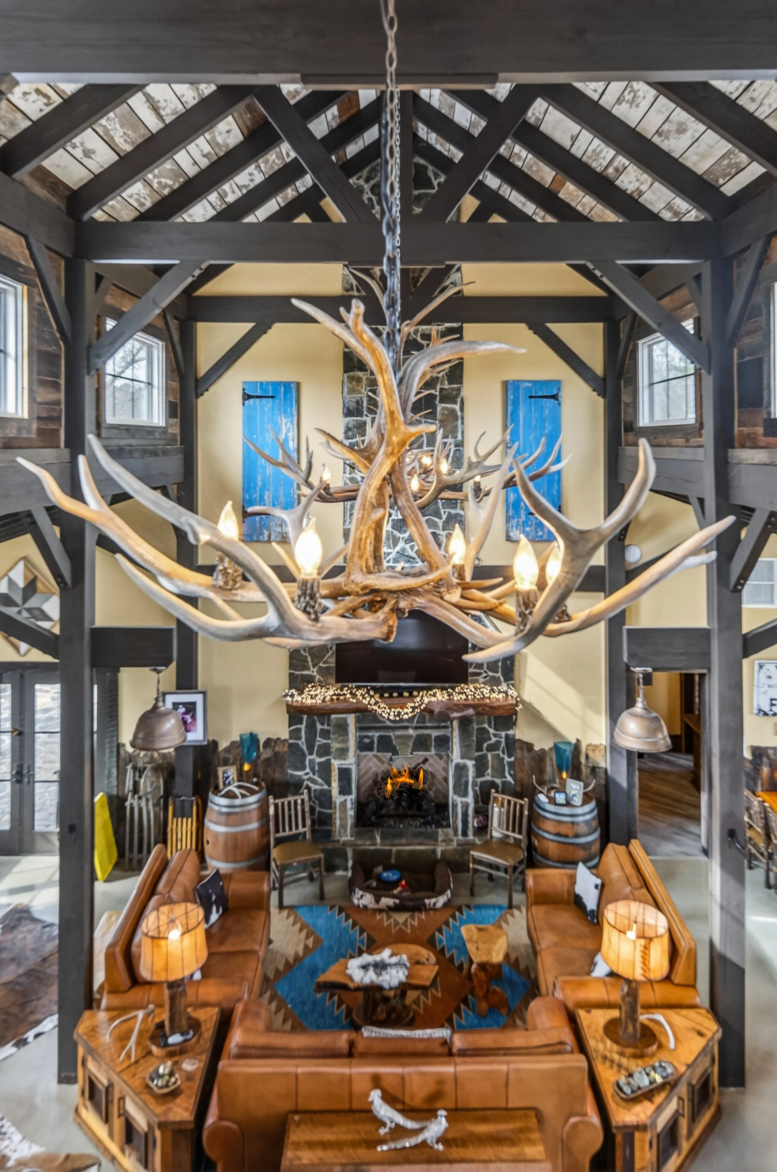 Interior of a rustic living room with a high ceiling, exposed beams, and windows. A large chandelier made of antlers hangs from the ceiling. There is a stone fireplace with decorations, surrounded by furniture including leather sofas, wooden tables, 