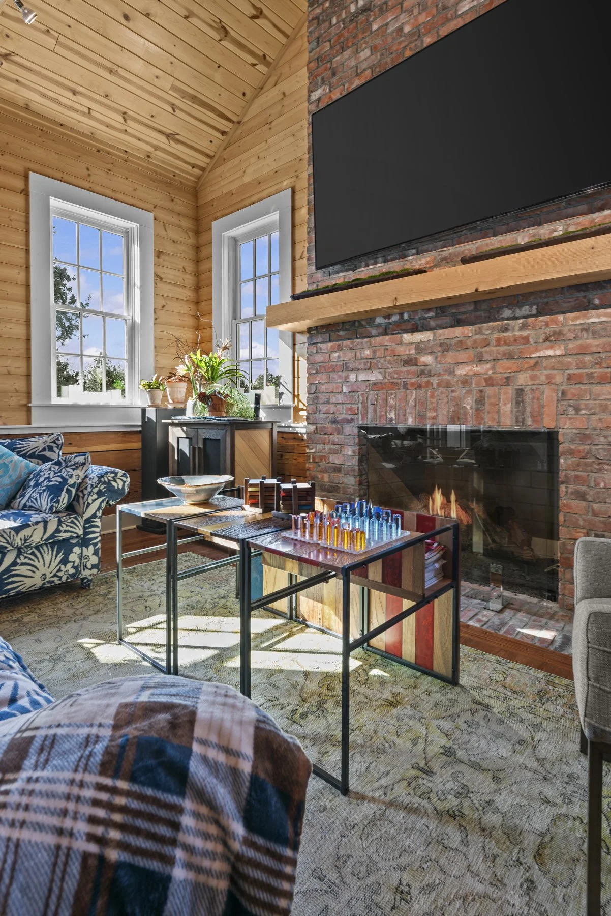 Living room with wood-paneled walls, brick fireplace, large windows, and a ceiling with wooden planks. There are houses visible outside through the windows. The room has a patterned couch, a colorful table with board games, and plants on a shelf by t