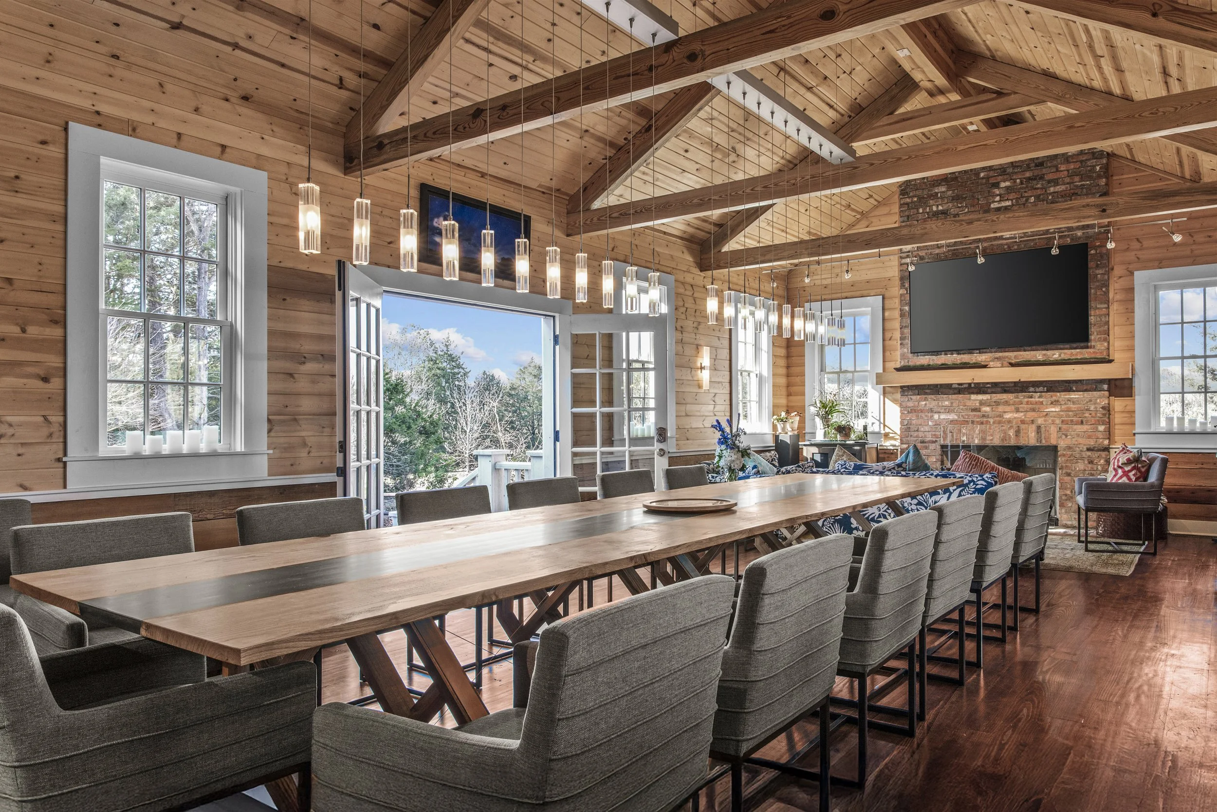 Rustic dining room with a long wooden table surrounded by gray upholstered chairs, large windows and open door leading outside, brick fireplace with a large flat-screen TV above, wood-paneled ceiling with exposed beams, pendant lighting, and scenic view outside.
