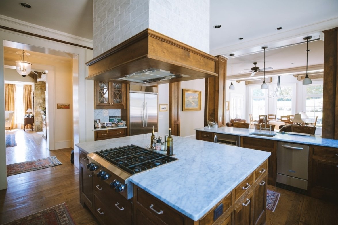 A modern kitchen with a marble island, wooden cabinetry, stainless steel refrigerator, and a view into a dining area with large windows and hanging pendant lights.