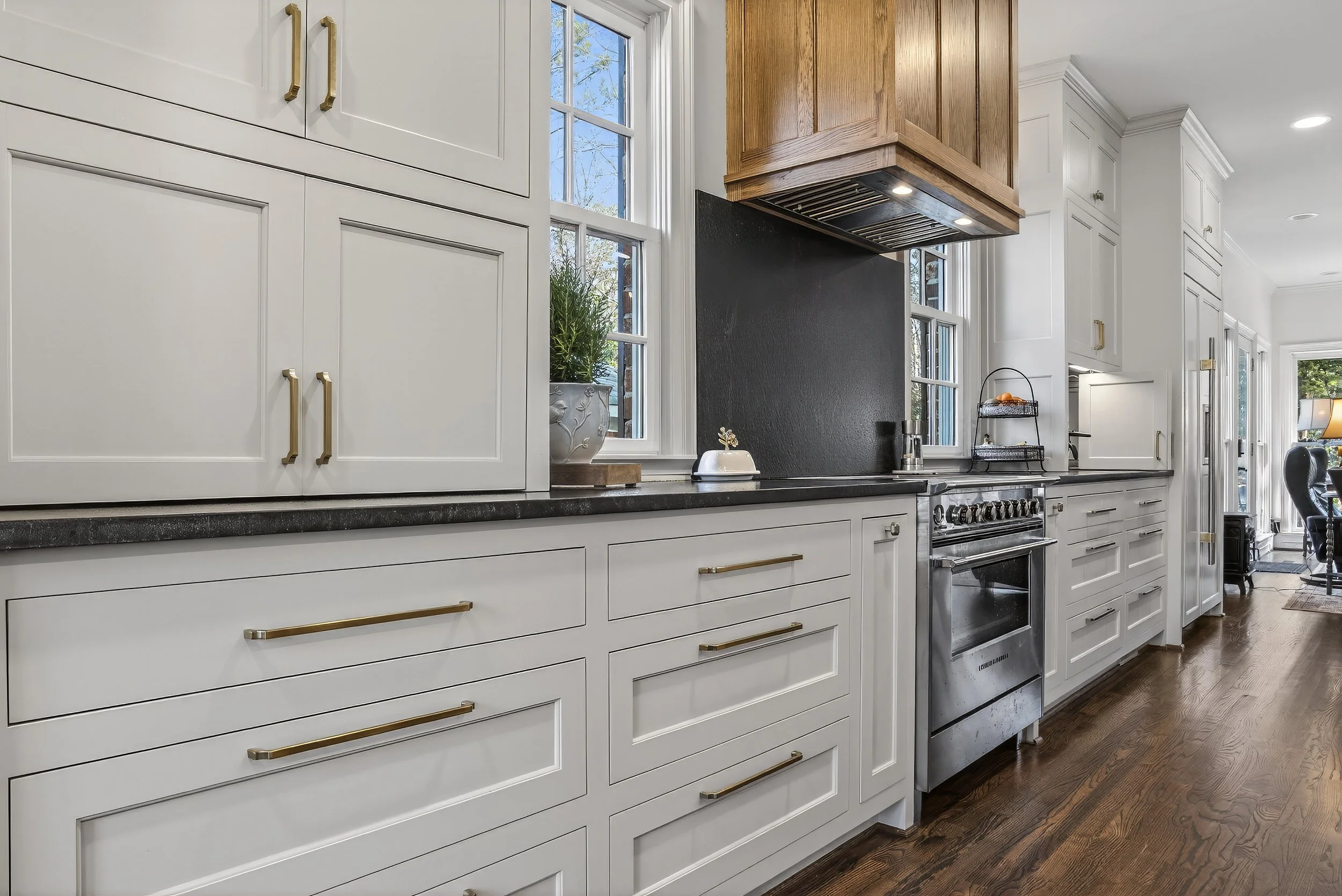 Modern kitchen with white cabinets, black countertop, stainless steel stove, wooden range hood, potted plant by window, and hardwood floor.