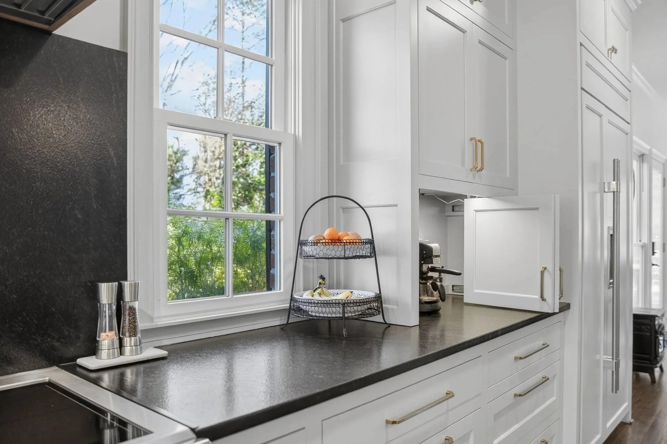 White kitchen counter with black countertop, window showing outdoor trees, two spice grinders, a two-tier wire fruit basket with oranges and bananas, a coffee machine, and white cabinets with gold handles.