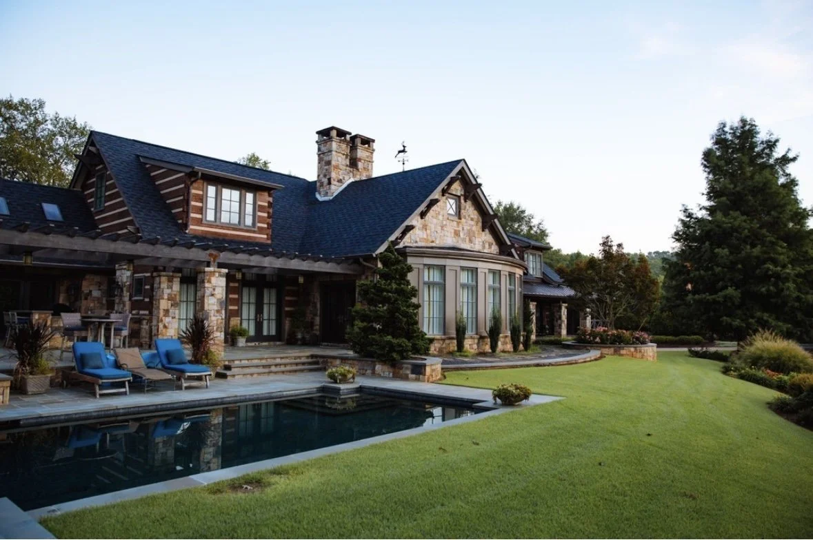 Exterior view of a large house with stone and wooden siding, a dark shingle roof, a swimming pool, and a well-maintained lawn and garden.