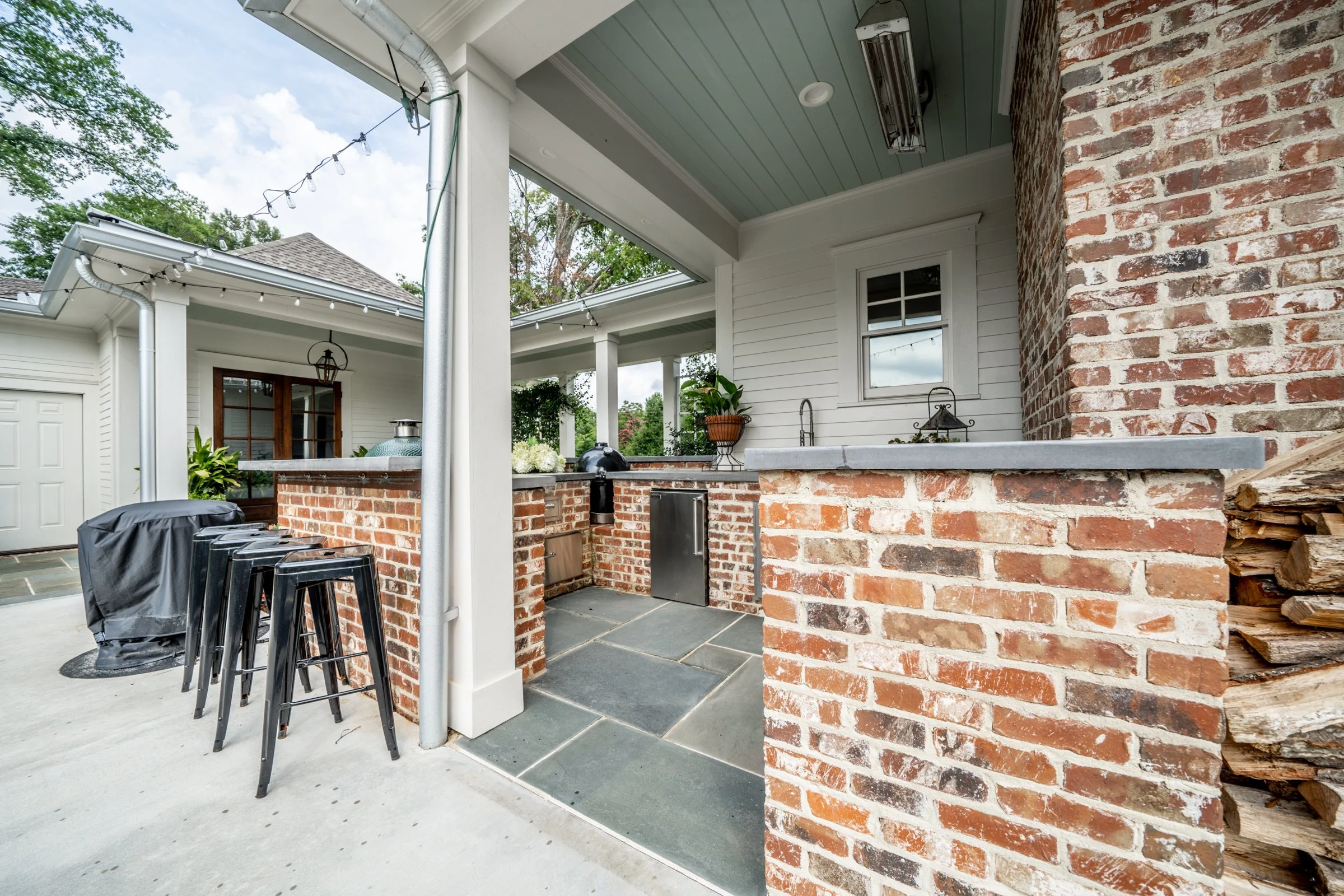 Outdoor kitchen area with brick and tile surfaces, a stainless steel countertop, a small window, potted plants, a covered grill, and stacked firewood, with string lights overhead.