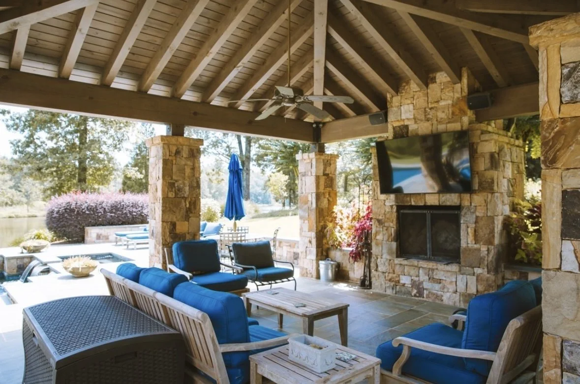 Outdoor patio with stone fireplace, blue cushioned chairs and sofas, a wooden coffee table, a wall-mounted flat-screen TV, and a ceiling fan under a wooden pavilion with a sloped roof. There is a view of a landscaped yard with trees and a patio umbre