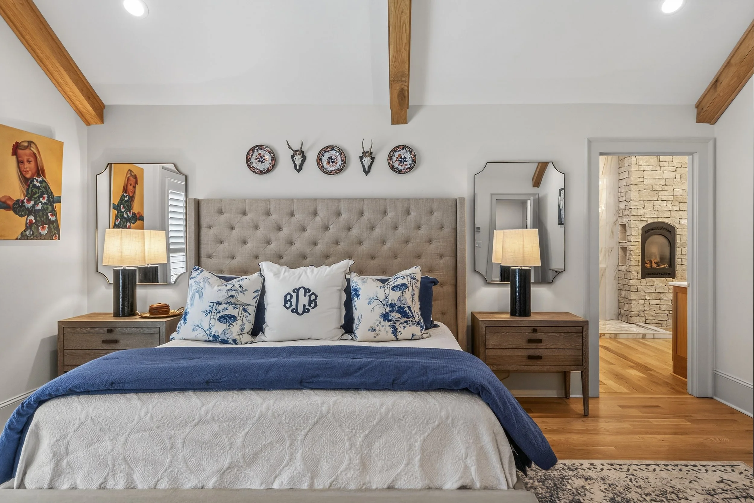 A bedroom with a large beige upholstered headboard, white bedding with blue accents, and decorative pillows including a monogrammed pillow. There are two wooden nightstands with matching lamps on either side of the bed. Above the headboard, there are