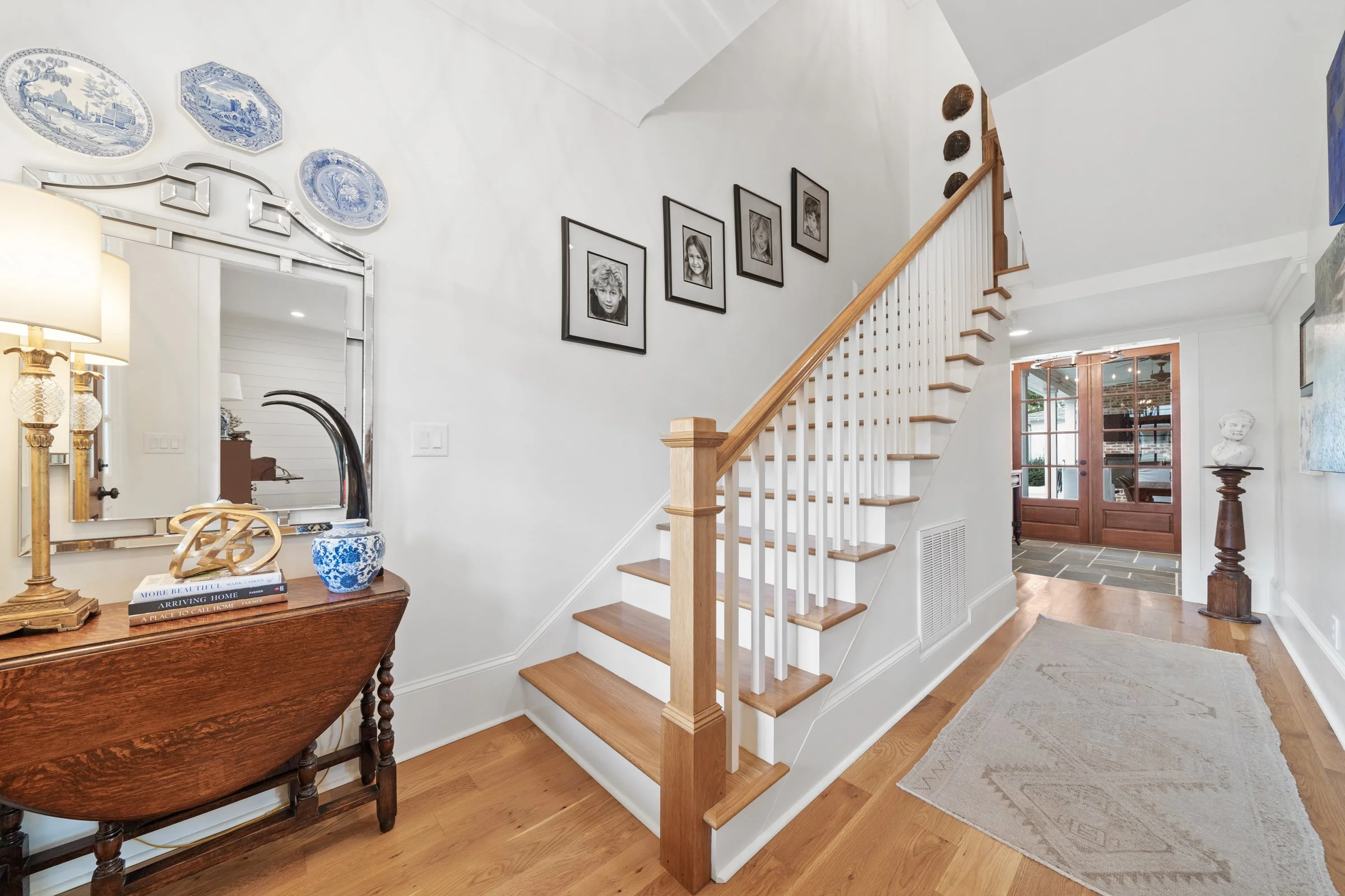 Entryway of a house with a staircase, decorative blue and white plates on the wall, framed black-and-white photos, and a wooden console table with books and decor.