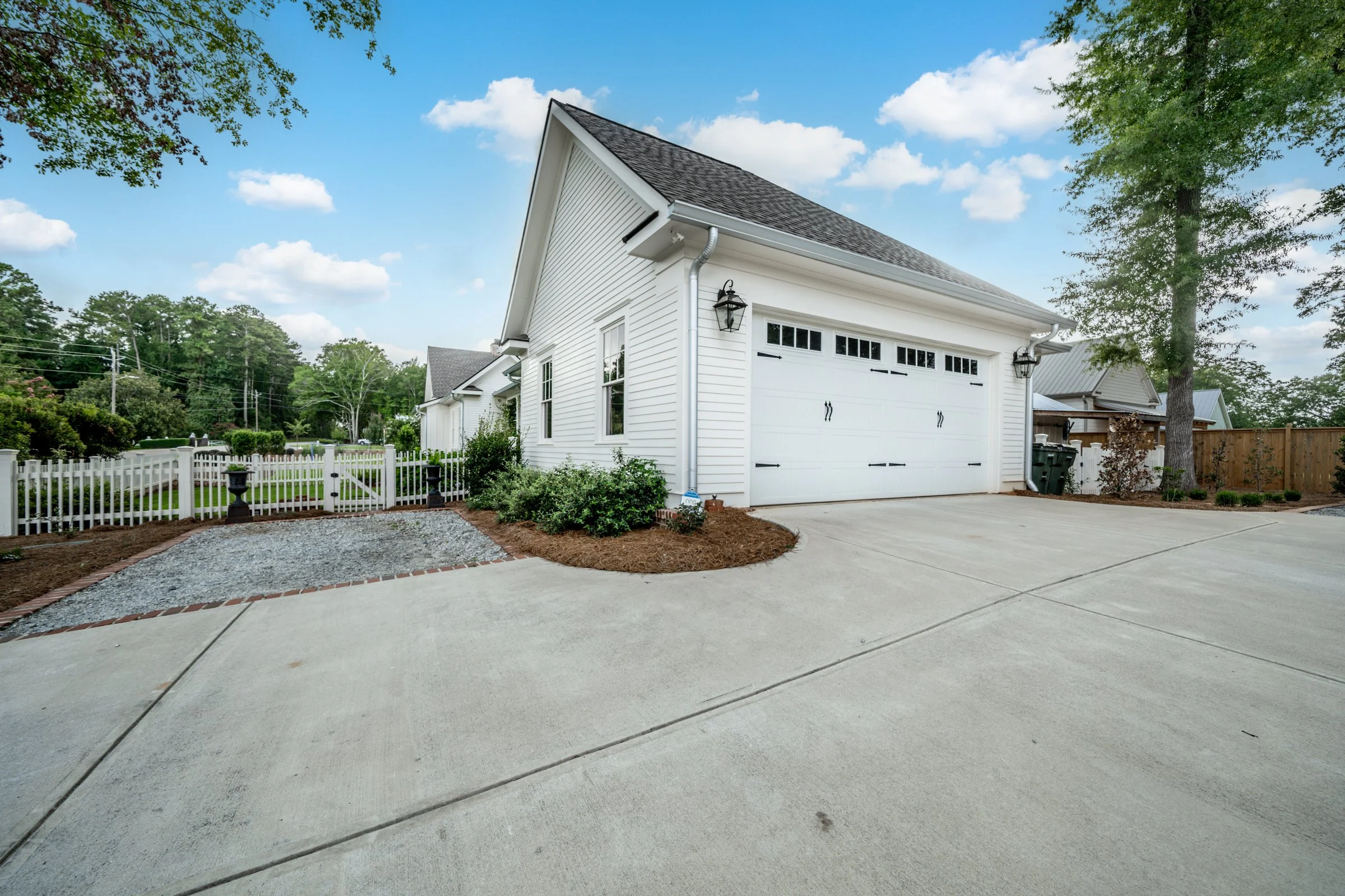 White house with attached garage, concrete driveway, small landscaped garden, mature trees, fenced yard, and blue sky with clouds.