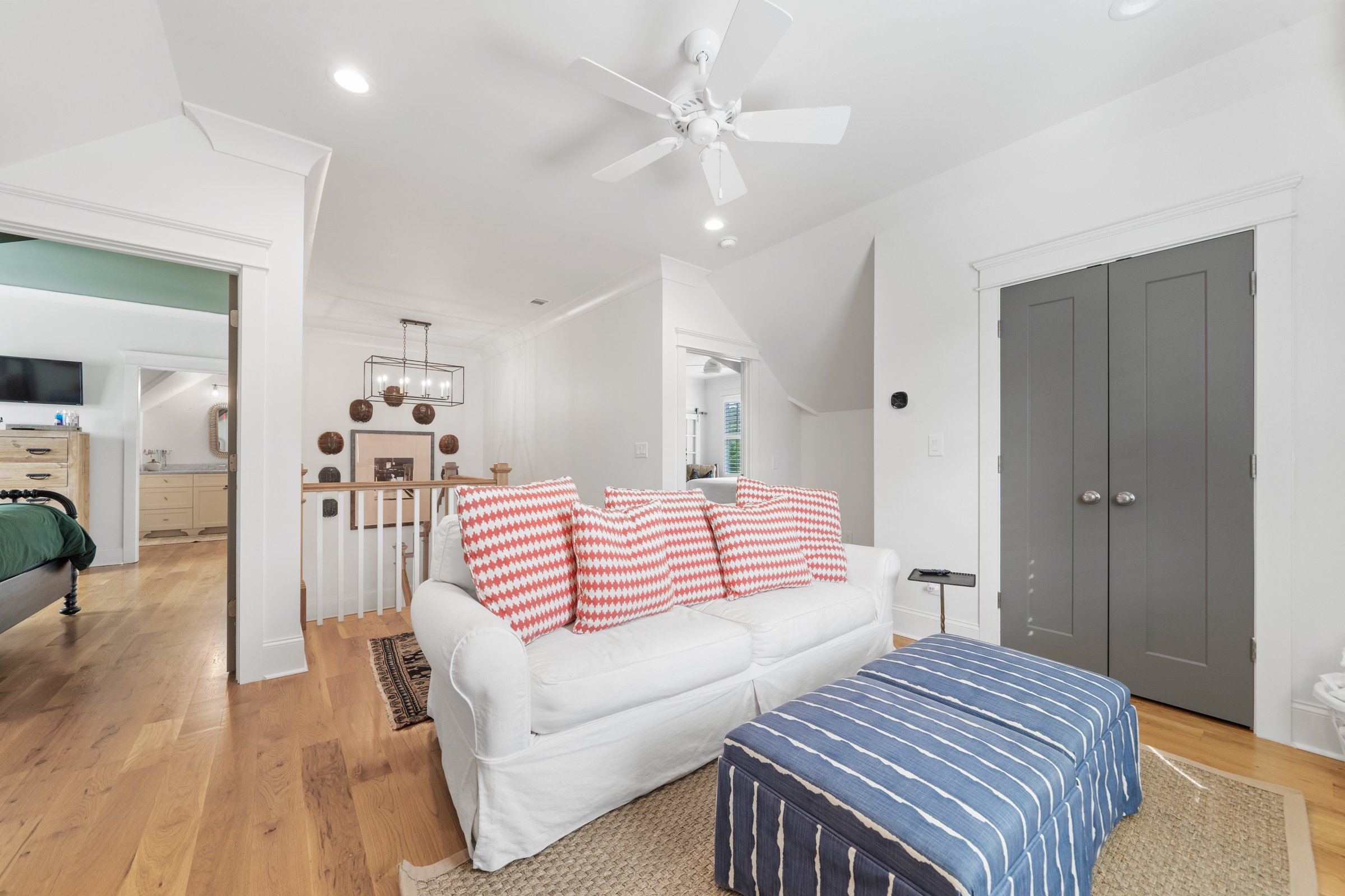 Living room with white sofa, striped blue ottoman, wood floor, built-in double closet with gray doors, ceiling fan, and an open doorway leading to other rooms.