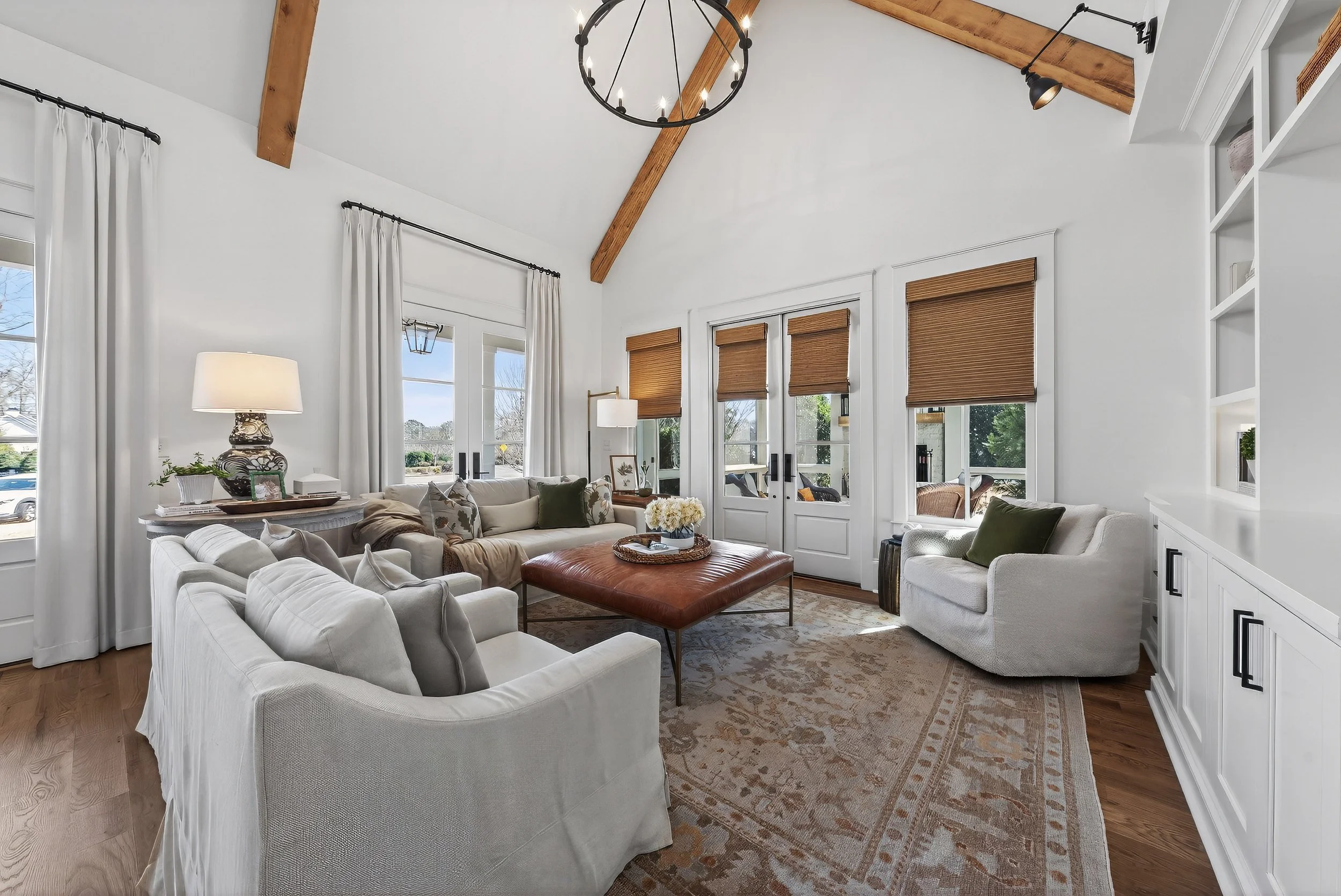 A spacious living room with white walls, large windows, and a high ceiling featuring wooden beams. The room is furnished with white sofas, an armchair, and a leather ottoman. There are curtains and wooden blinds on the windows, a rug on the hardwood 