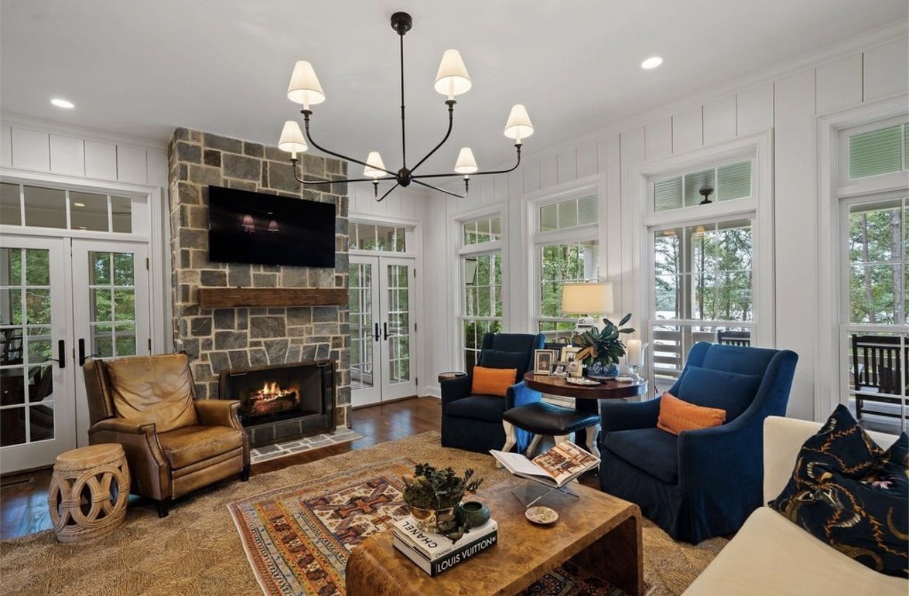 Living room with a stone fireplace, multiple large windows, and blue and brown furniture, including armchairs and a white sofa, with a wooden coffee table and a chandelier.