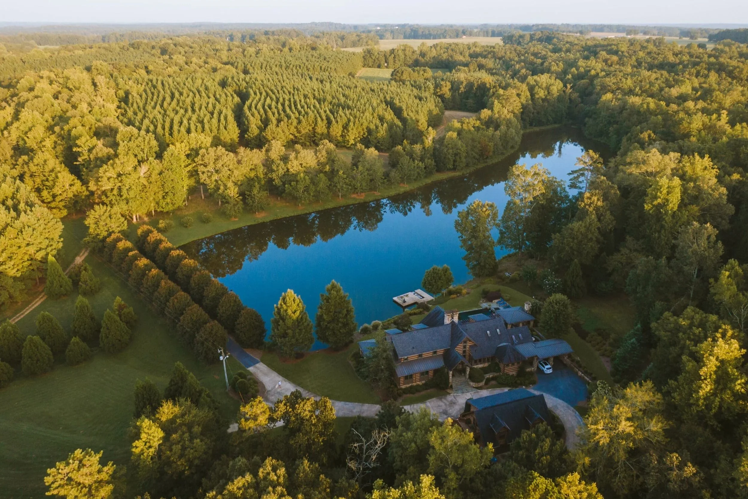 An aerial view of a large house with a pool, surrounded by a lush landscape including a pond, trees, and fields on a clear day.