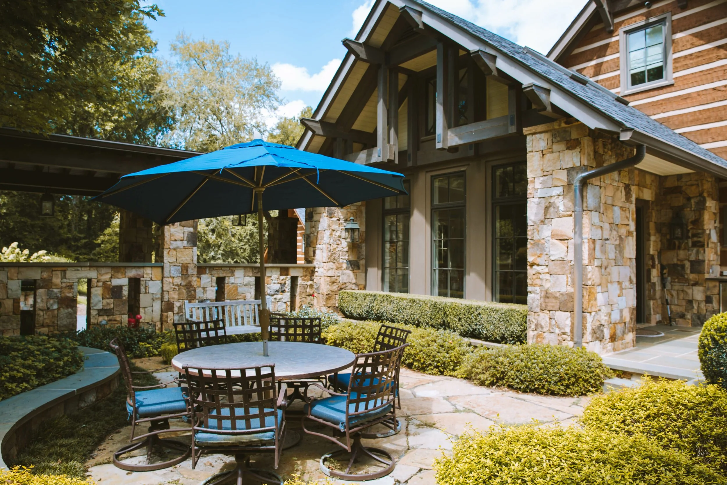 A backyard patio with a round table, six chairs with cushions, and a large blue umbrella. The background features a house with stone and wood siding, large windows, and a sloped roof. There are green bushes, shrubs, and trees surrounding the patio on