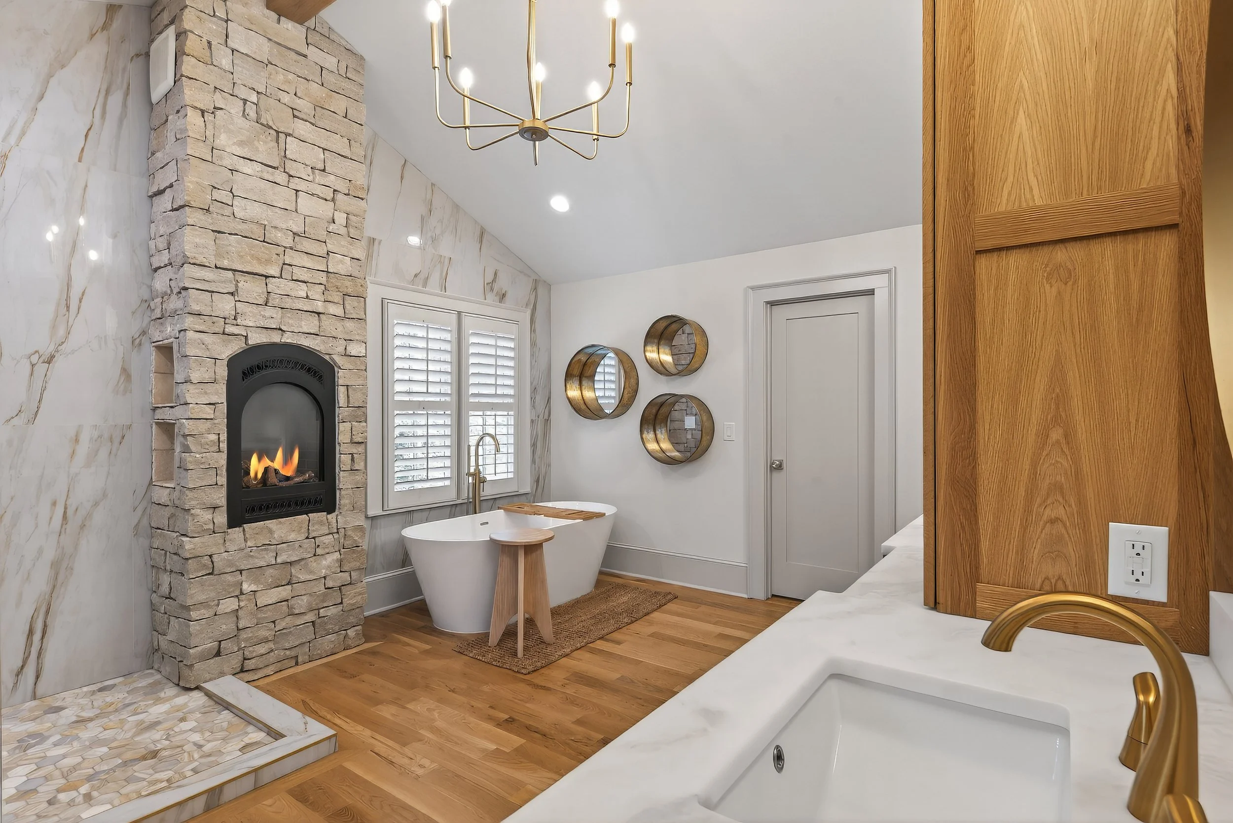 A modern bathroom with a stone fireplace, a bathtub with a wooden stand, wooden and metallic decor, and a large mirror with wooden framing.
