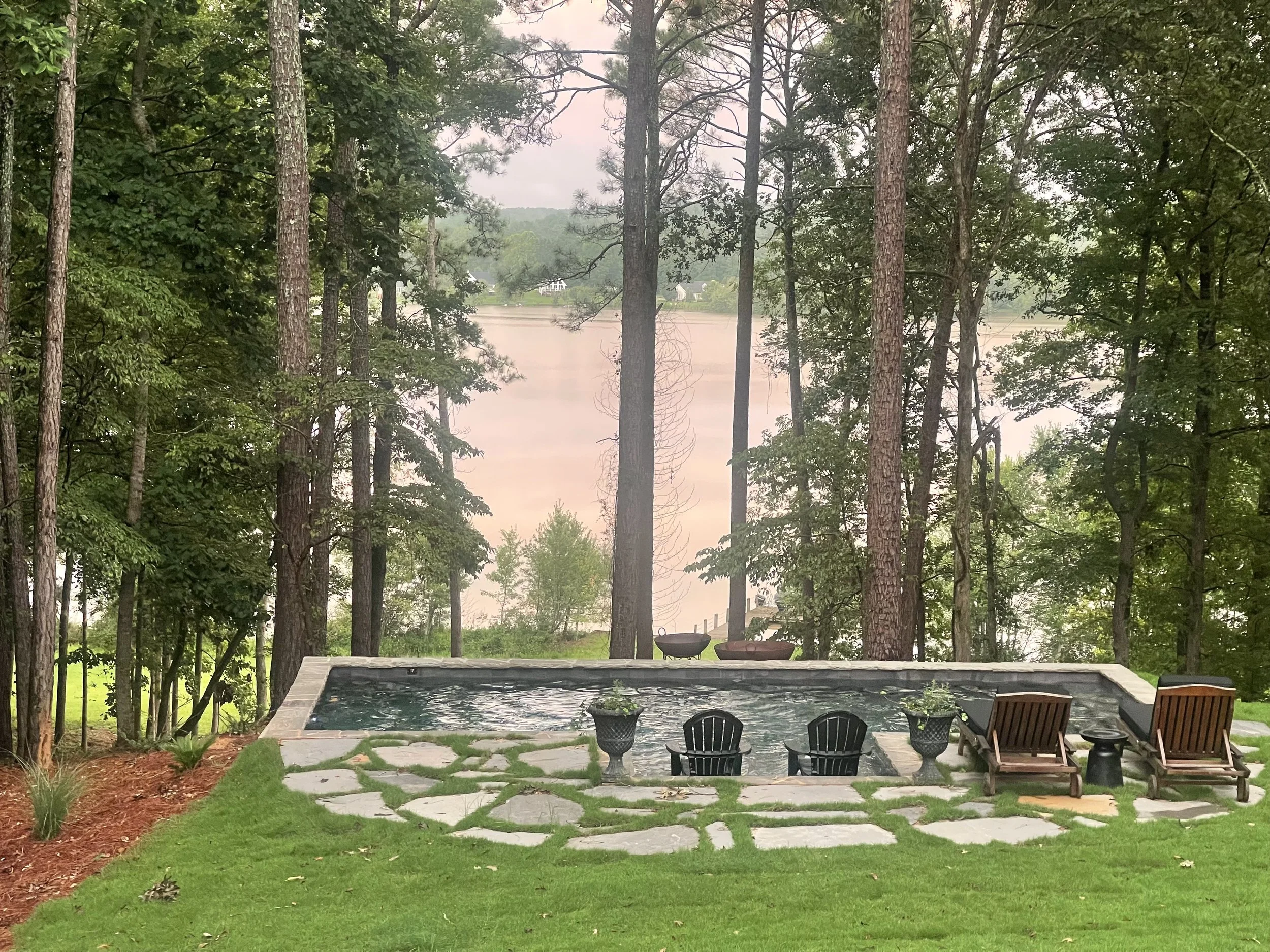 A backyard with a small pool, surrounded by stone pavers and green grass, overlooking a lake with trees and a cloudy sky in the background.