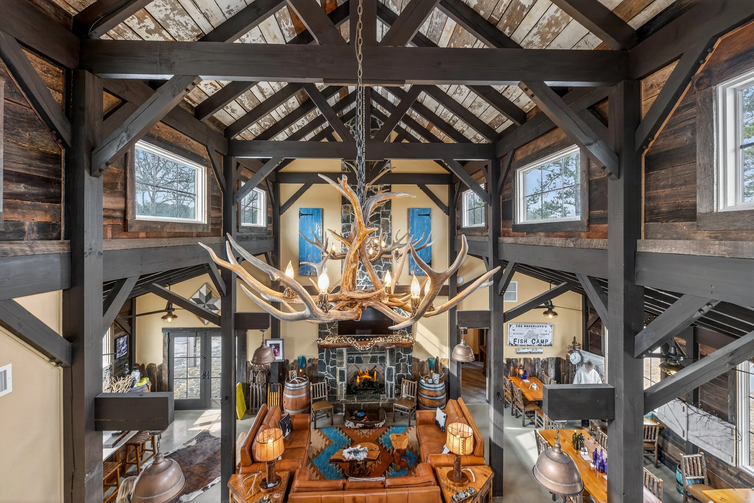 A rustic living room with high vaulted ceiling, central fireplace, and large windows. A chandelier made of antlers hangs from the ceiling. Leather sofas and wooden furniture are arranged around a stone fireplace.
