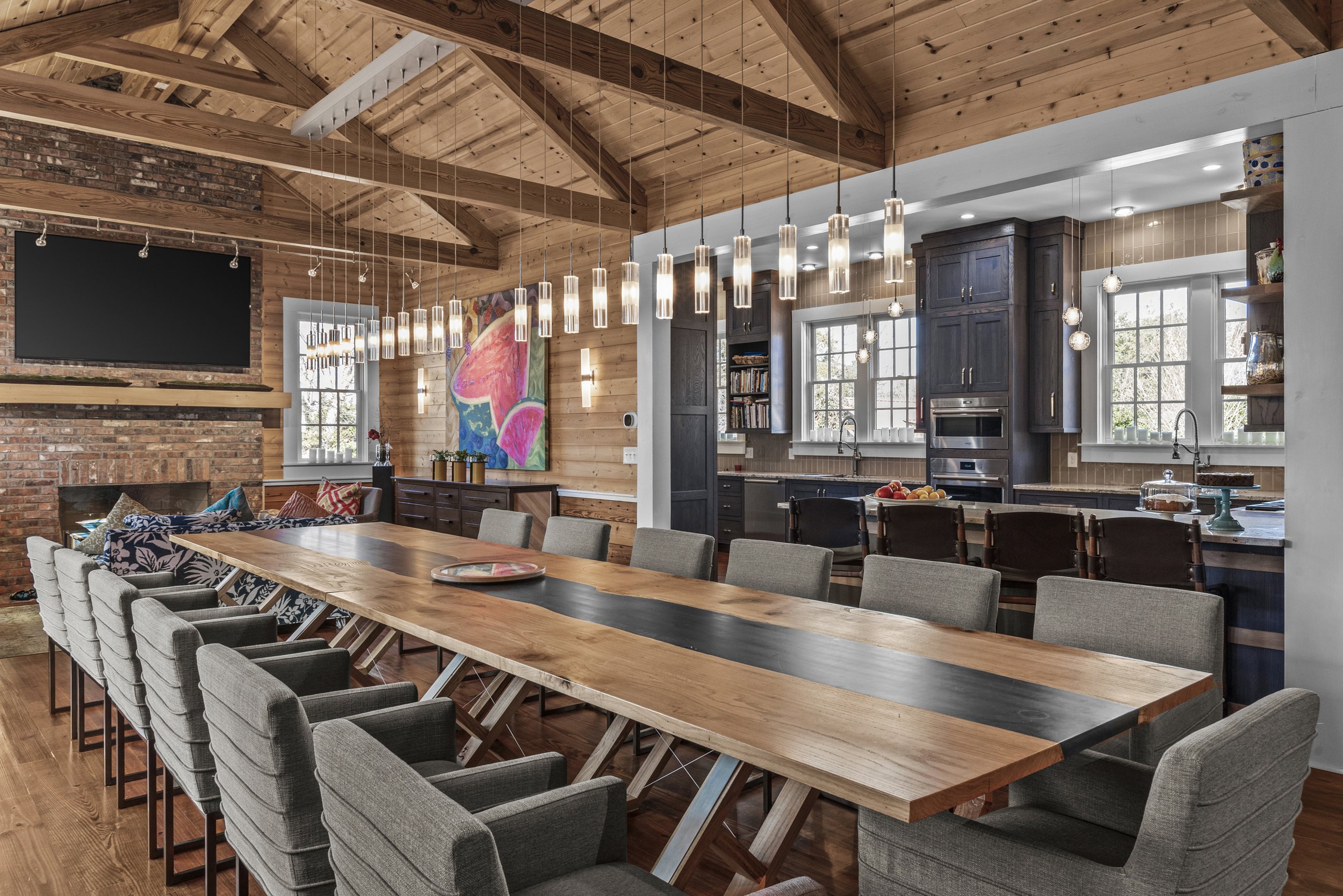 Open-concept kitchen and dining area with a large wooden table, gray chairs, a brick fireplace, wood ceiling, colorful artwork, and multiple hanging pendant lights.