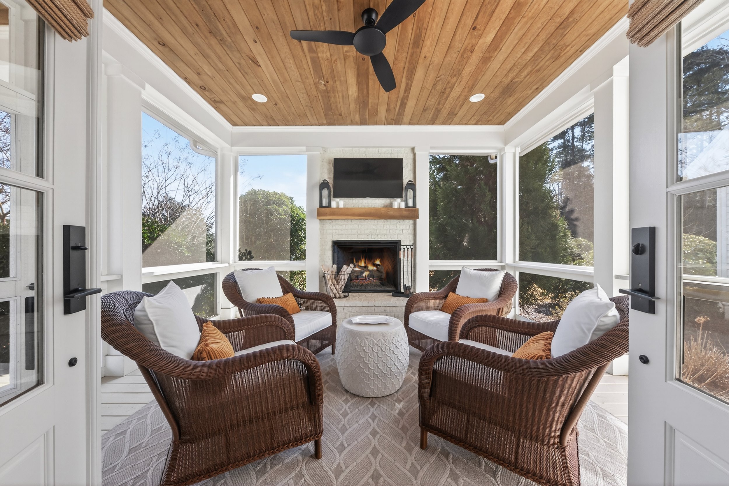 Sunlit screened-in porch with wicker chairs, white cushions and throw pillows, a white fireplace with a TV mounted above, and a wooden mantel, surrounded by windows showing trees outside.