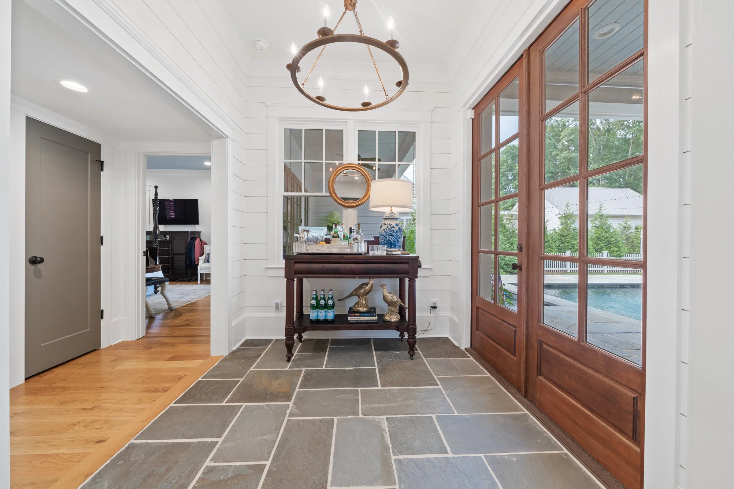 Entryway with a wooden side table, decorated with two bird sculptures, bottles of sparkling water, and a lamp, with large glass-paneled wooden doors leading outside to a pool area. A window behind the table reflects part of the interior, and a chande