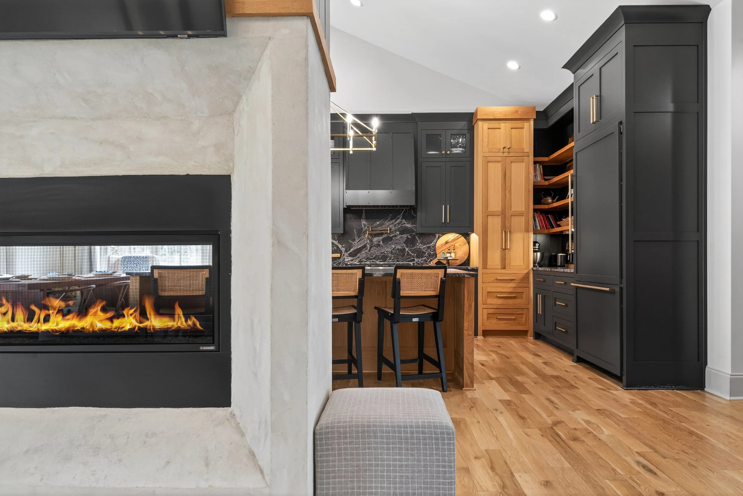 Modern kitchen with black and wood cabinetry, black marble backsplash, and barstools, with a partial view of a fireplace in the foreground.