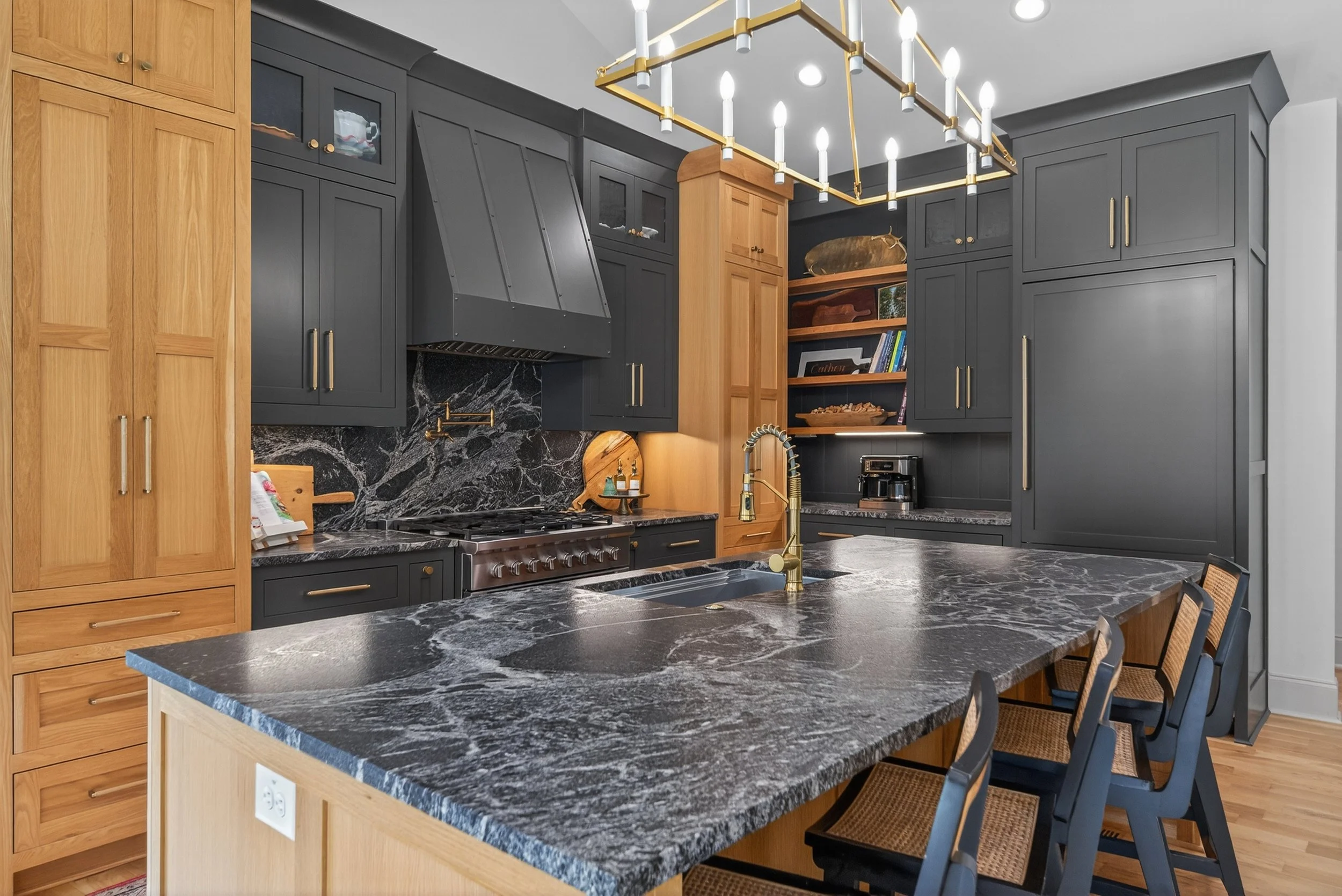 Modern kitchen with dark gray cabinets, black marble countertops, and a kitchen island with seating. Includes a stainless steel stove, a black range hood, gold fixtures, open shelving with books, and a large gold chandelier.