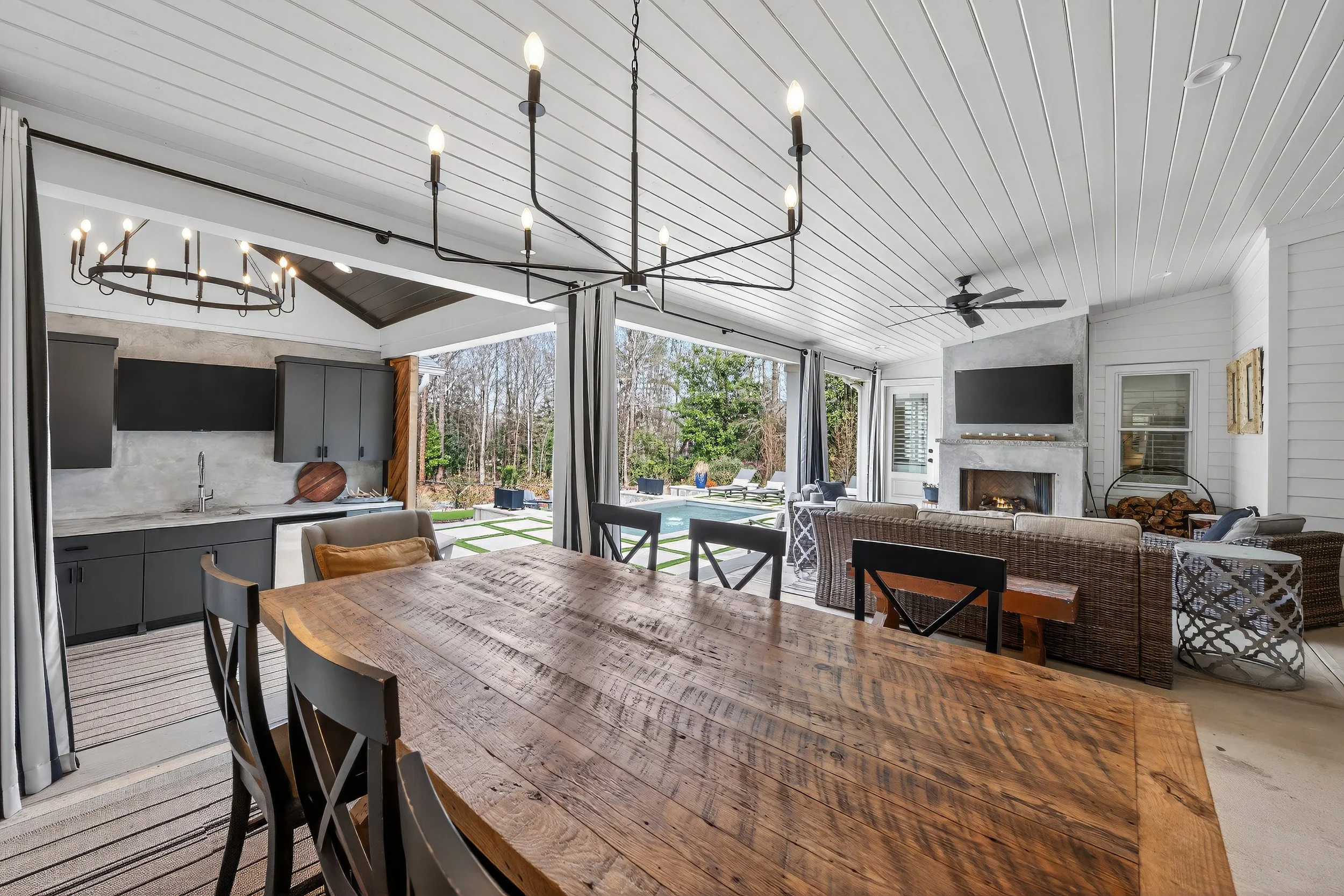 Open-concept living and dining area with view of outdoor pool and backyard, featuring a wooden dining table, black and wicker seating, modern lighting fixtures, white paneling ceiling, a fireplace, and large windows with curtains.