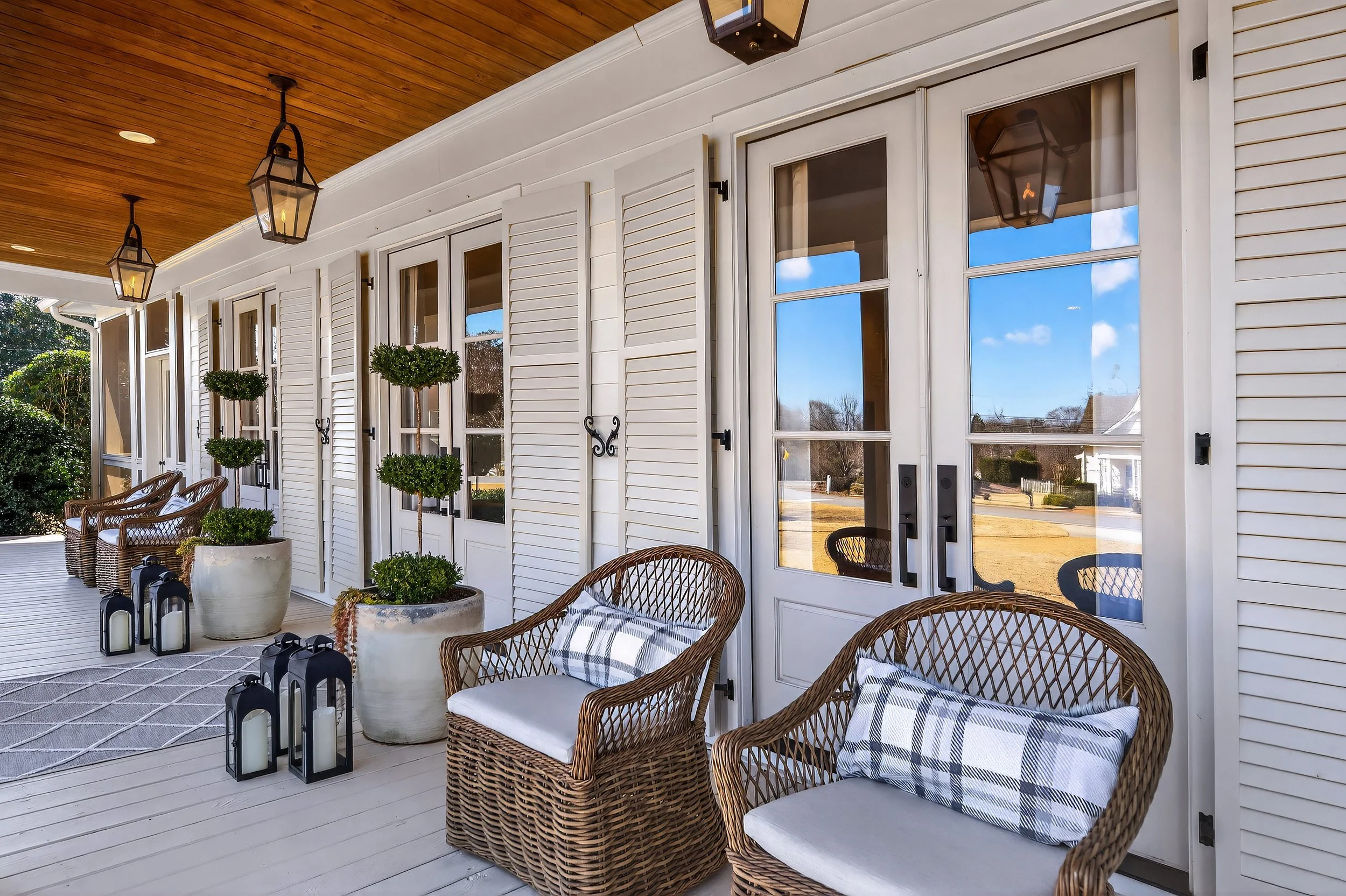 Front porch of a house with outdoor wicker chairs, potted trees, and lanterns, set against a white wall with large glass doors.