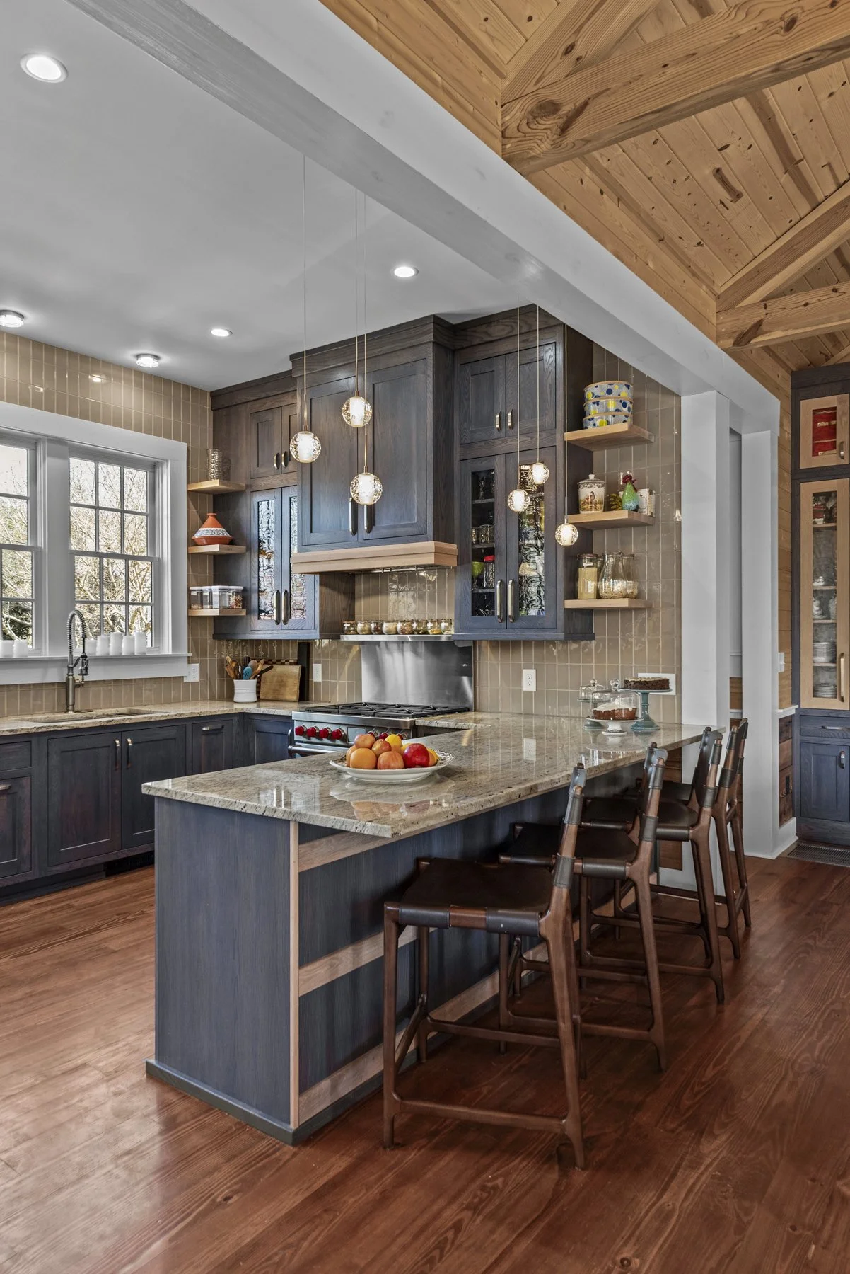 A modern kitchen with dark wood cabinets, beige tiled walls, a granite island with fruit, a large window, pendant lights, and wooden ceiling beams.