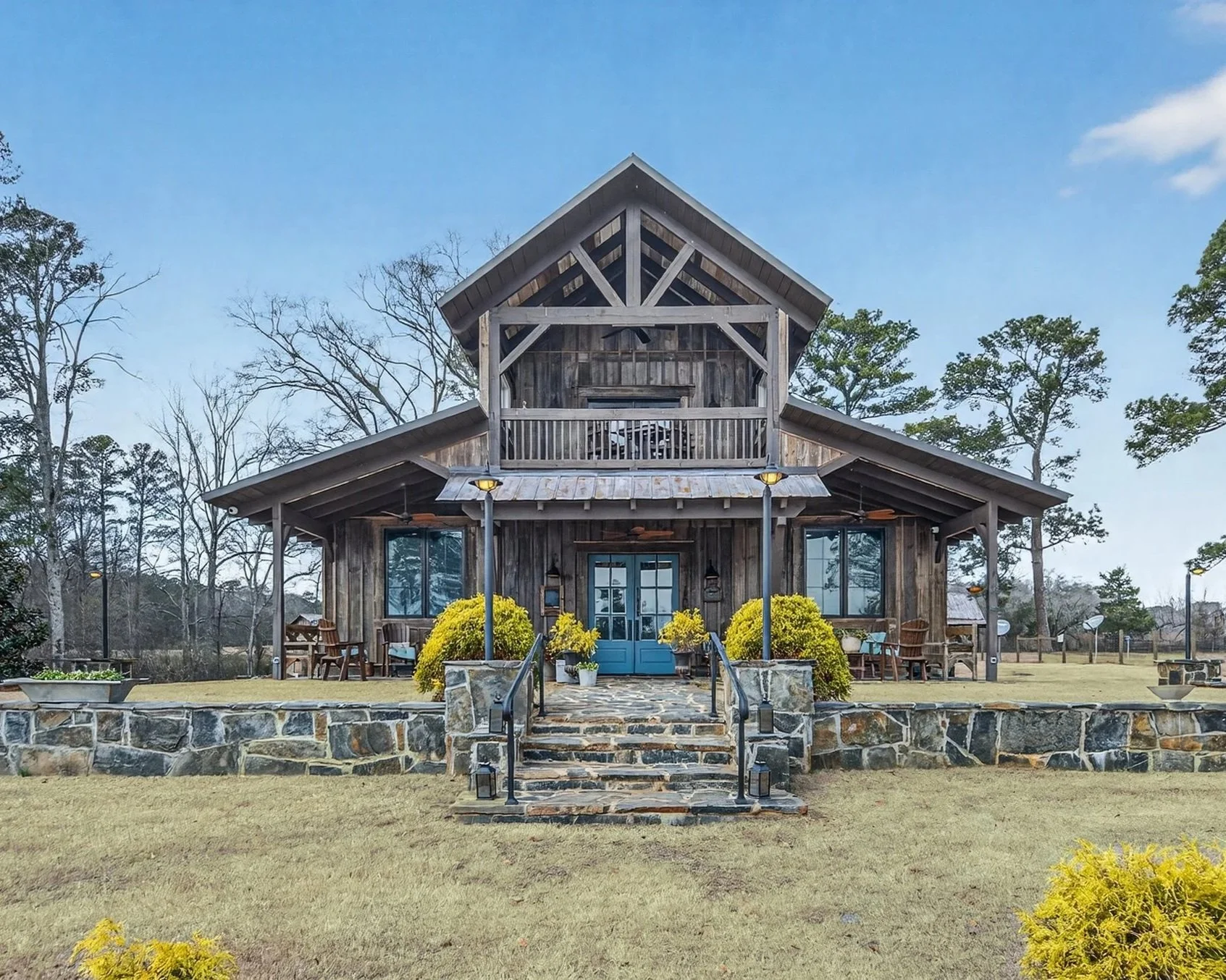 A two-story rustic wooden house with a stone staircase leading to a blue front door, surrounded by yellow bushes and outdoor seating, set in a grassy yard with trees in the background under a partly cloudy sky.