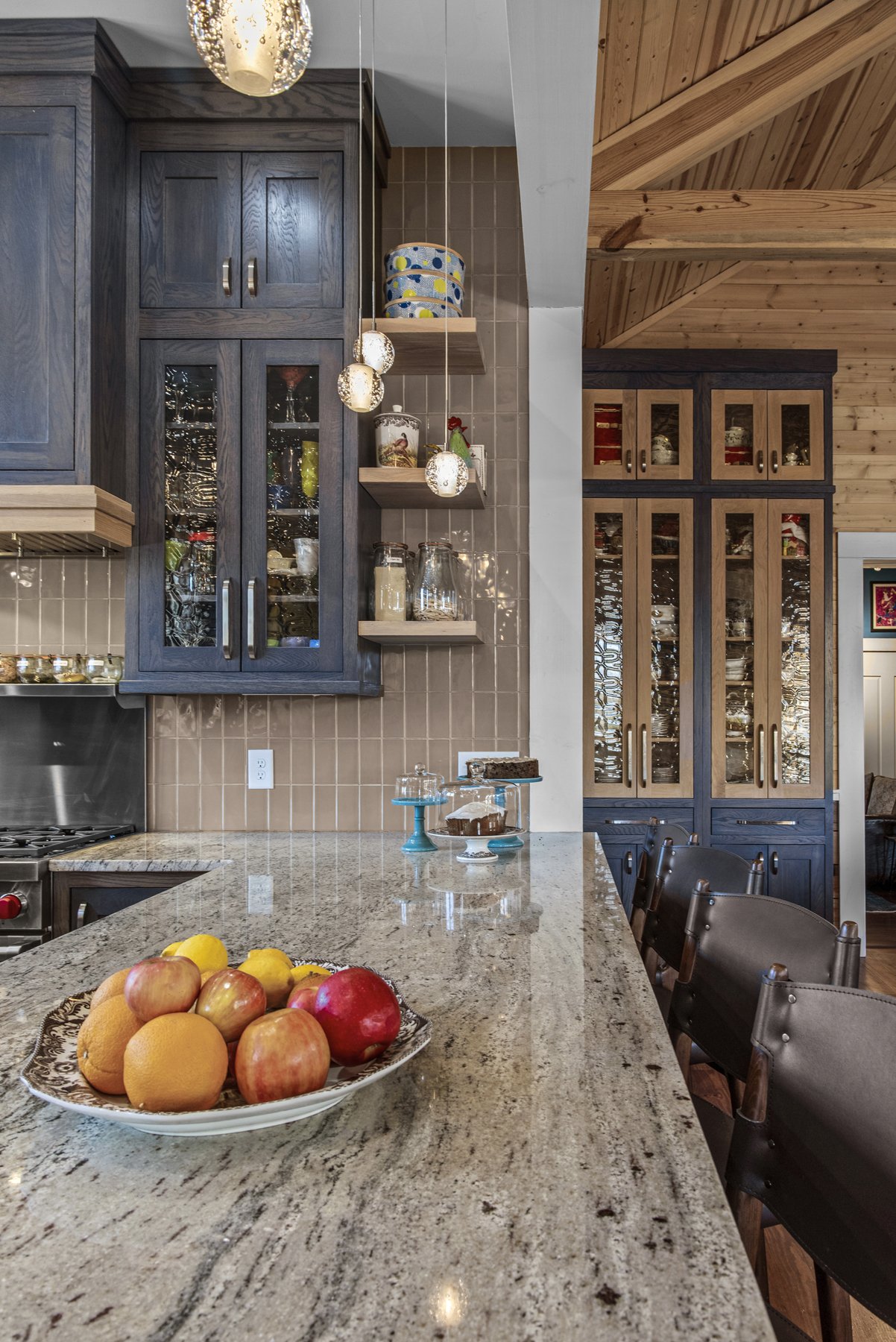 A kitchen with dark and light wood cabinets, a granite countertop, and a stone tile backsplash. There's a bowl of assorted fruits on the counter, hanging pendant lights, and open shelves with jars and decorative items.