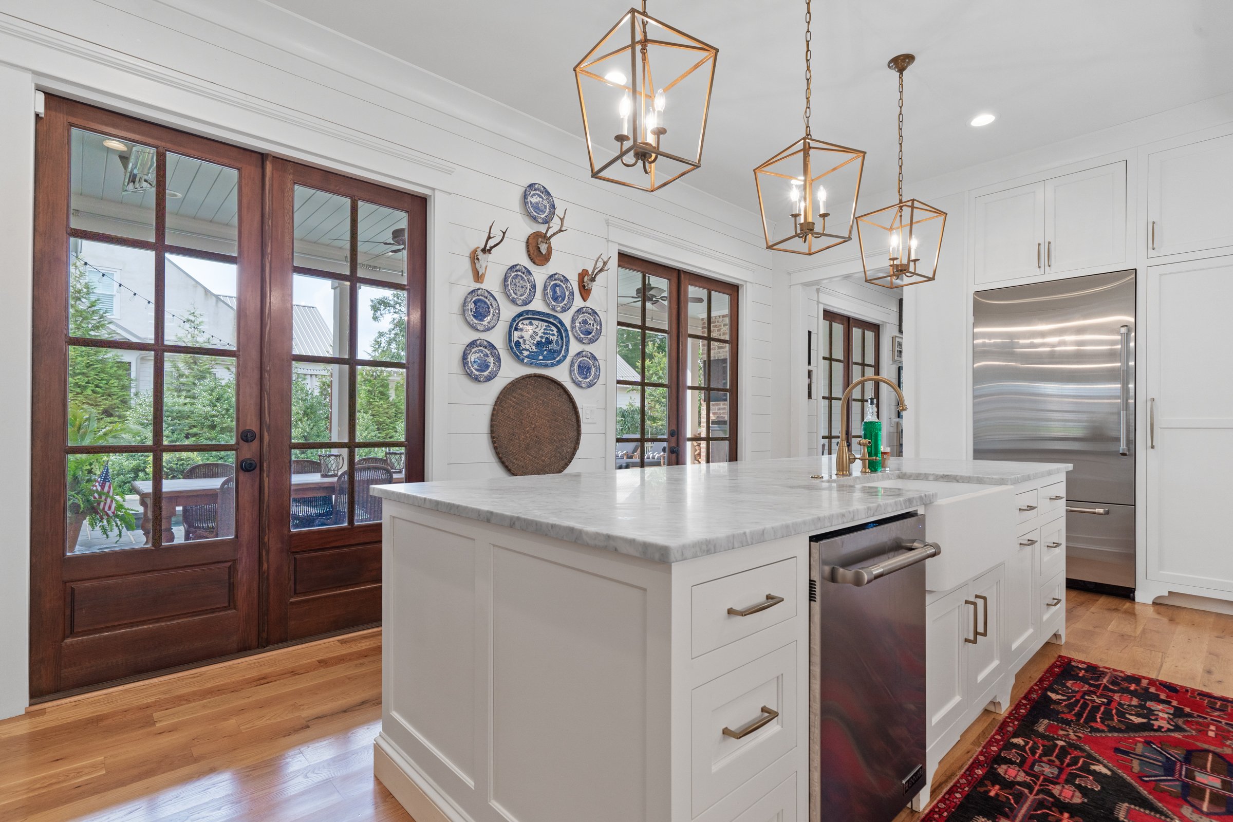 Kitchen with white cabinets, marble island, hardwood floors, and wooden double doors with glass panes leading outside. Decor includes blue and white plates and deer antler wall art. Three pendant lights hang over the island, and a stainless steel ref